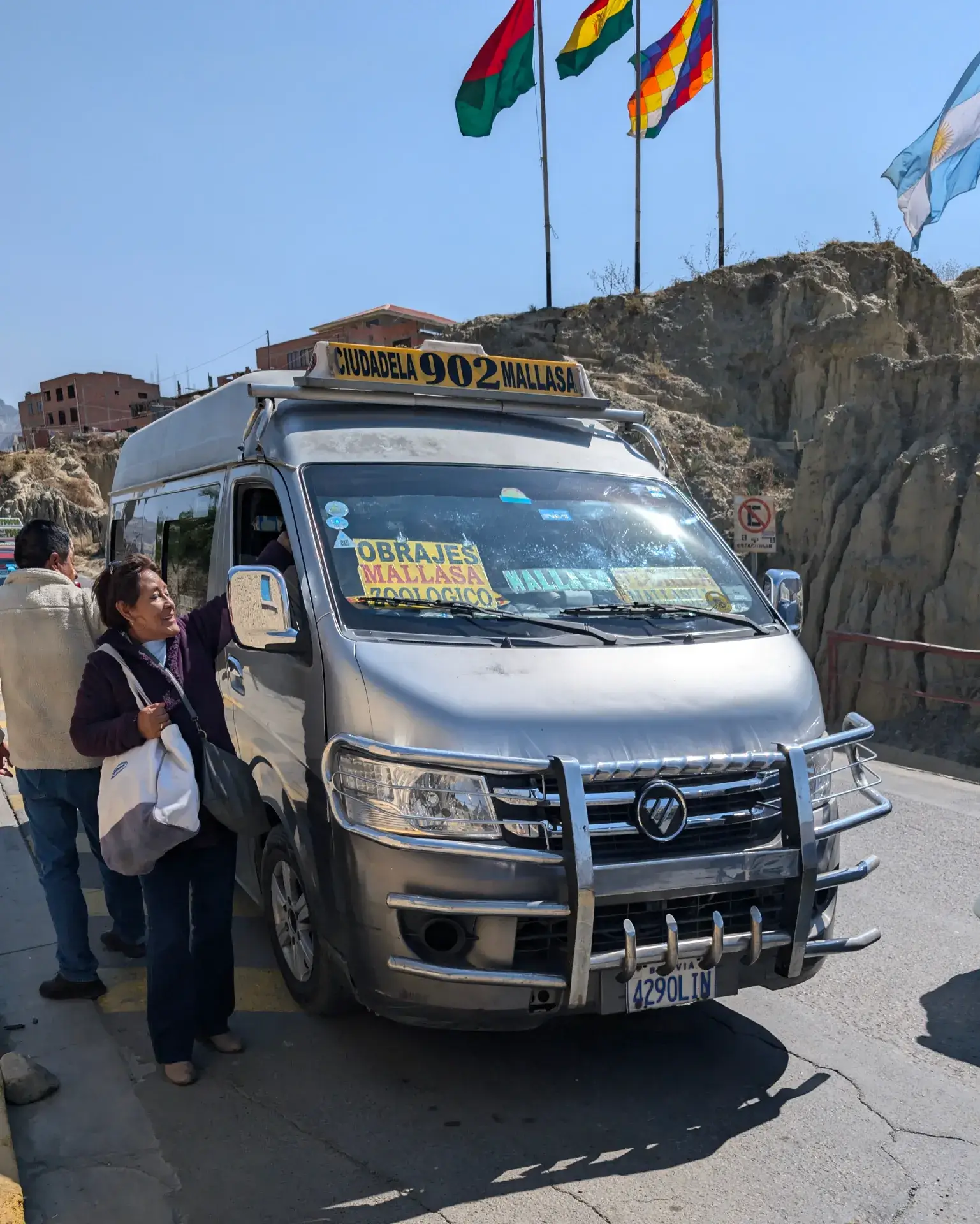 ラパスの険しい坂道や複雑な路地を網羅する、市民の足「トゥルフィ（Trufi）」。フロントガラスには「OBRAJES」「MALLASA」といった目的地が手書きの看板で掲げられ、乗り降りの合間に交わされる人々の活気が伝わってきます。頭上にはボリビア国旗や多民族国旗（ウィパラ）がはためき、乾燥した土色の崖が背景に迫るこの光景は、まさに標高4,000m級の摩天楼ラパスならでは。現地の生活のペースに身を任せ、未知のエリアへと踏み出す高揚感に包まれる、旅の移動のワンシーンです。