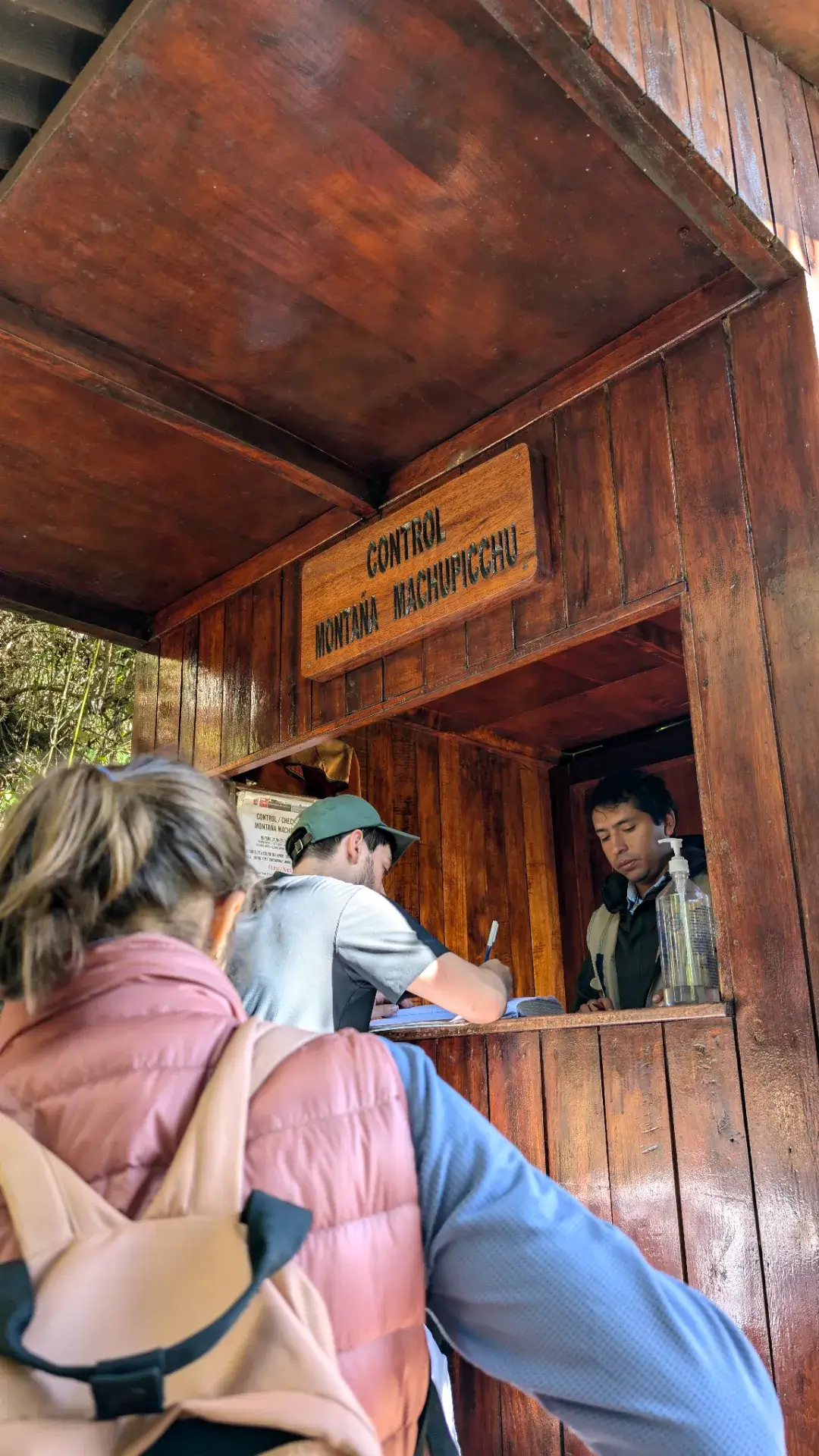 マチュピチュ遺跡を一望できる絶景ポイント「マチュピチュ山（Montaña Machu Picchu）」への登山口。木造の簡素なコントロールポイントでは、登山者の安全管理のために名前や入山時刻の記帳が行われます。順番を待つ旅行者の背中からは、これから始まる急勾配の石段への緊張感と、頂上で待つ絶景への期待が伝わってきます。遺跡のさらに高みを目指す、本格的なアドベンチャーの始まりを告げる重要な一コマです。