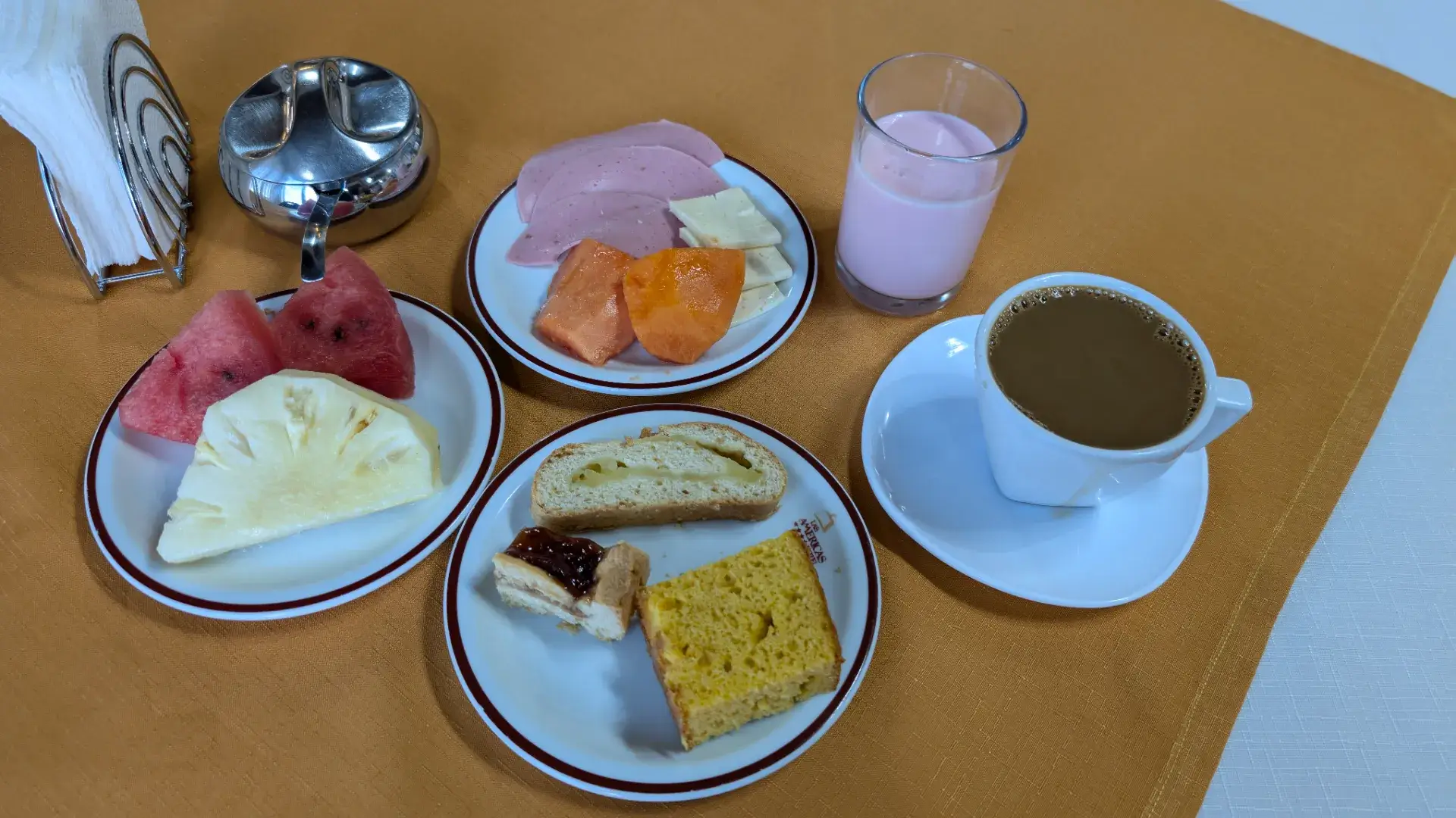 ボリビア、サンタ・クルスのホテルで提供された朝食。スイカやパイナップル、パパイヤなどの新鮮なフルーツの盛り合わせ、ハムとチーズ、パン、そしてカップに注がれたコーヒーとピンク色のスムージーがオレンジ色のテーブルクロスの上に並んでいる。