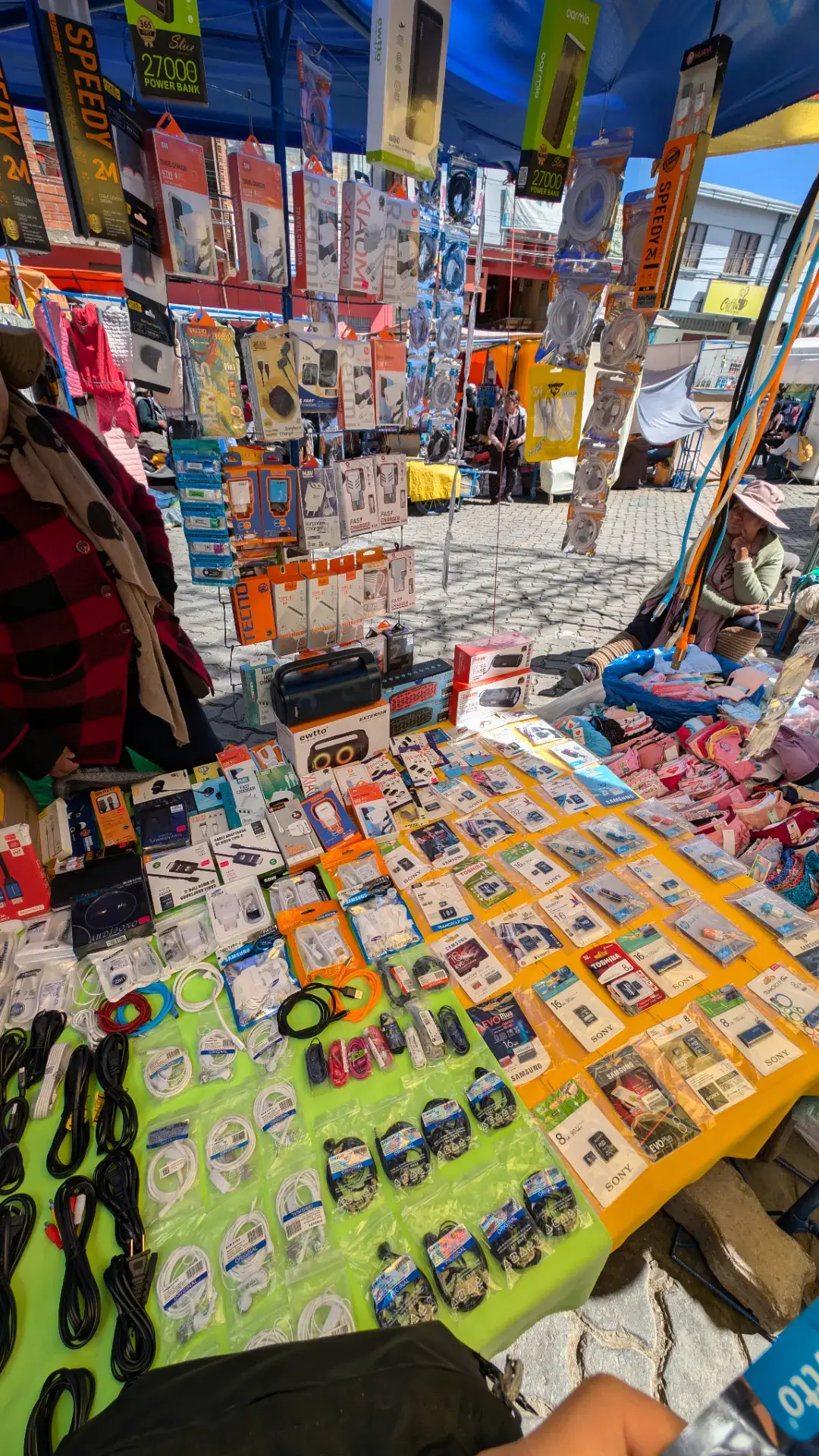 ラパスの路上に突如現れる、ハイテクと日常が混在するマーケット。黄色いクロスが敷かれたテーブルには、SDカードやイヤホン、USBアダプタなどの小さなガジェット類がパズルのように隙間なく並べられています。頭上にはモバイルバッテリーやケーブルが吊るされ、その奥では地元の店主たちが静かに客を待つ、ラパス特有の商景。最新のデジタル製品が、アンデスの乾いた空気と素朴な市場の喧騒の中に溶け込んでいる、ボリビアの「今」を象徴する光景です。