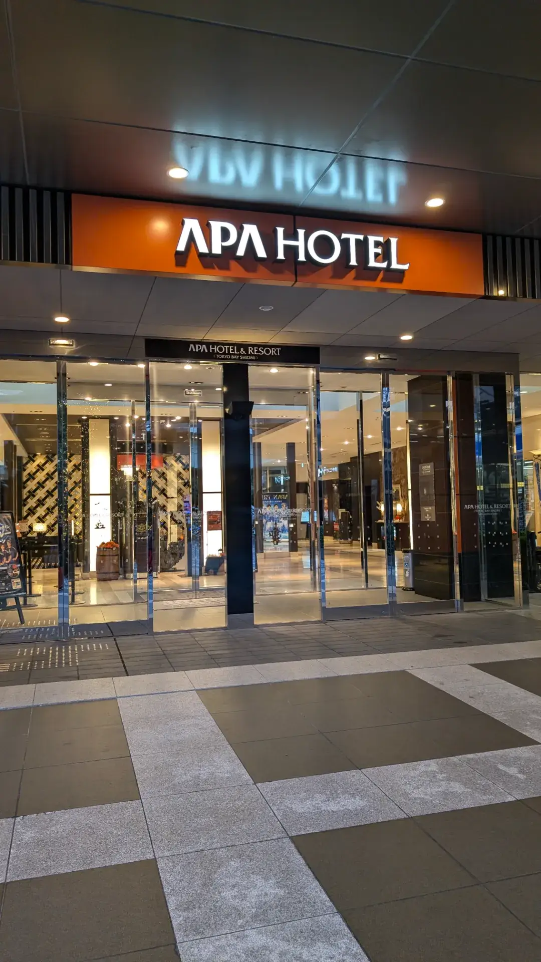 夜のアパホテル&リゾート〈東京ベイ潮見〉のエントランス。明るくライトアップされたオレンジ色の「APA HOTEL」の看板と、ガラス張りの開放的なロビーへと続く入り口の風景。