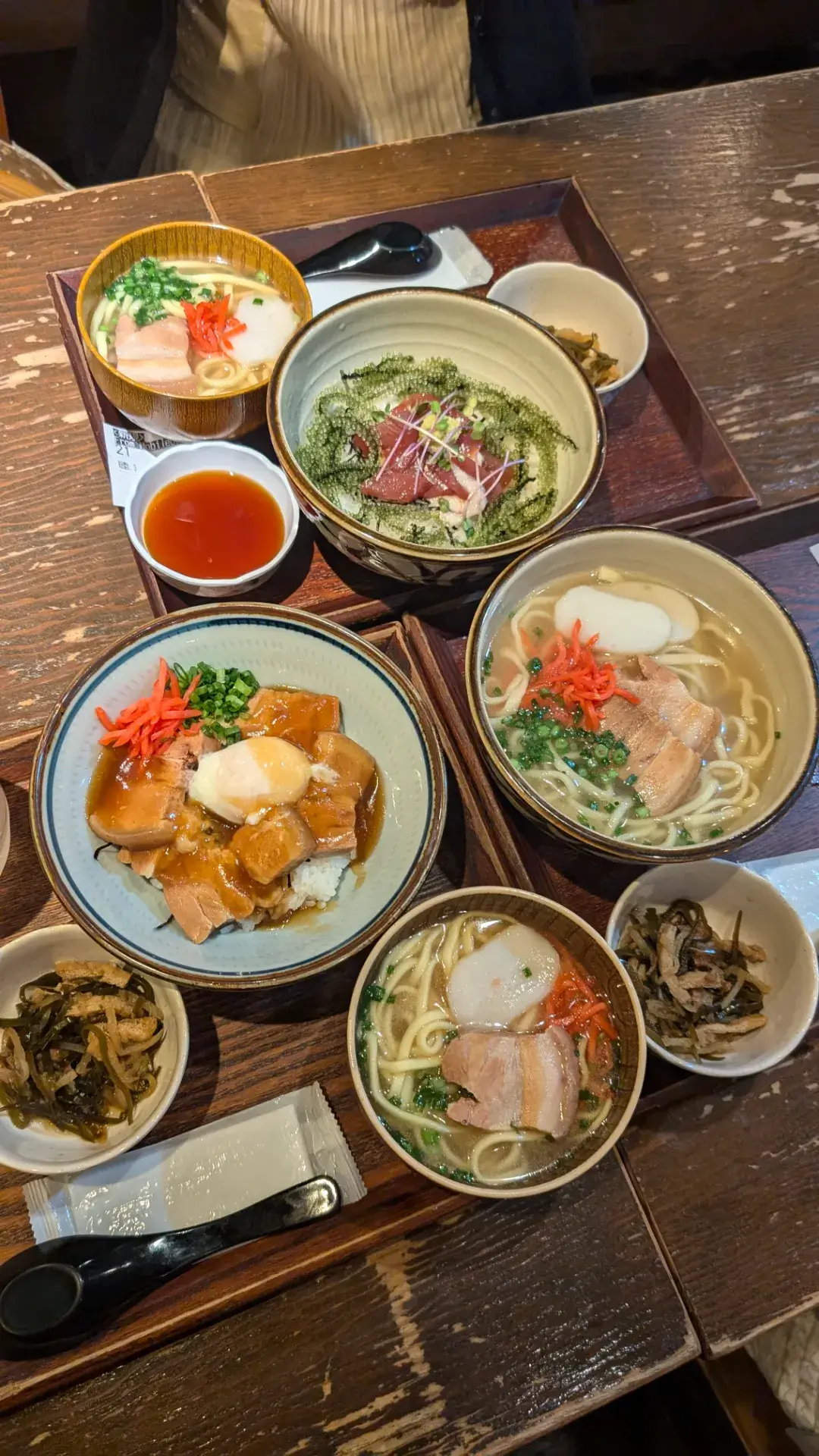 沖縄料理店「海人酒房」でのランチ風景。ソーキそば、海ぶどうとマグロの丼、ラフテー丼(温泉卵のせ)など、ボリューム満点のセットメニューが木製トレイに並んでいる。紅生姜や小鉢の彩りが鮮やかな会食の様子。