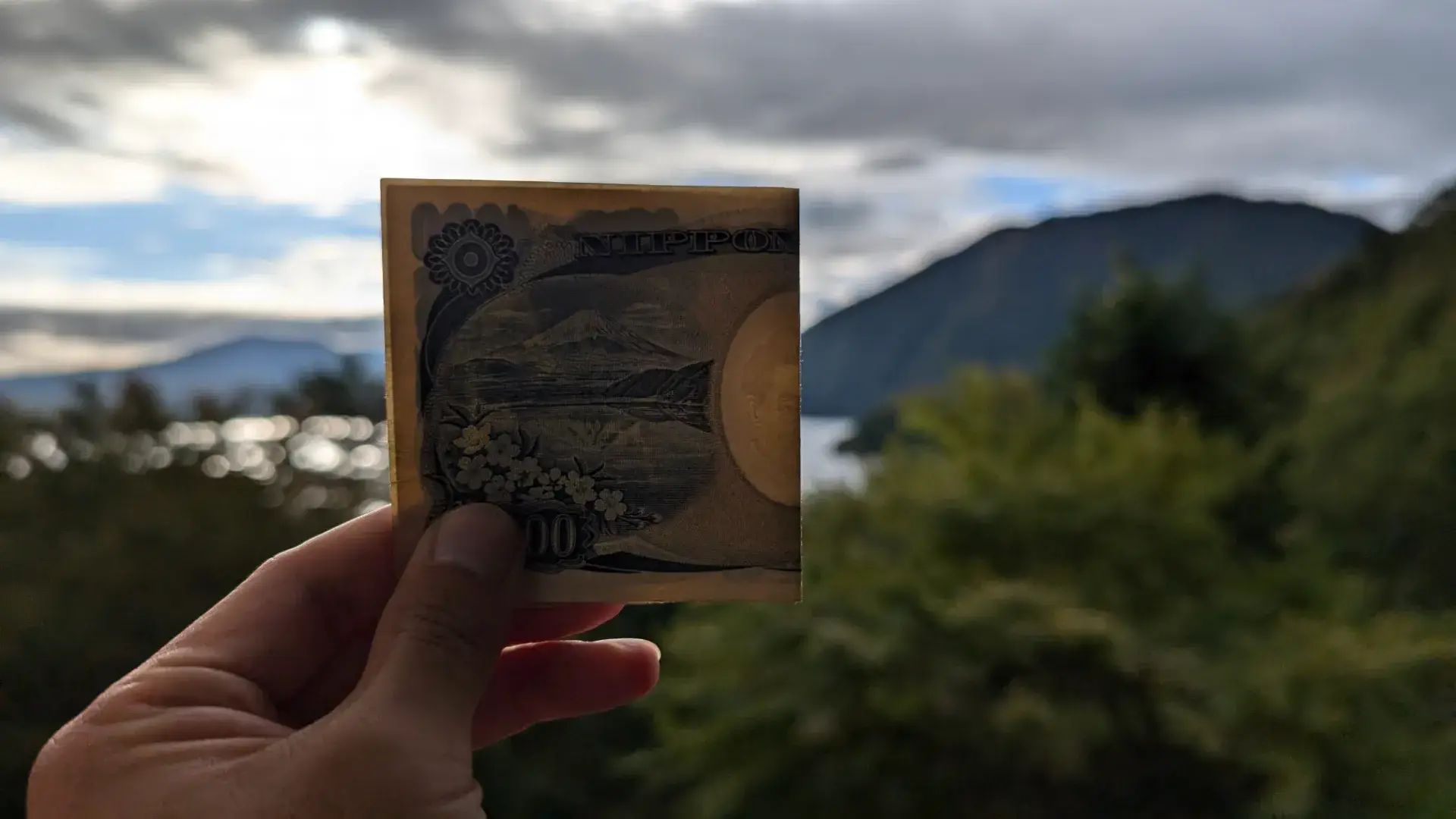本栖湖の風景をバックに、千円紙幣の裏面に描かれた富士山と本栖湖の図案を重ねて比較している様子。実際の景色と紙幣のデザインを一致させる、観光客に人気のフォトスポットでの1枚。