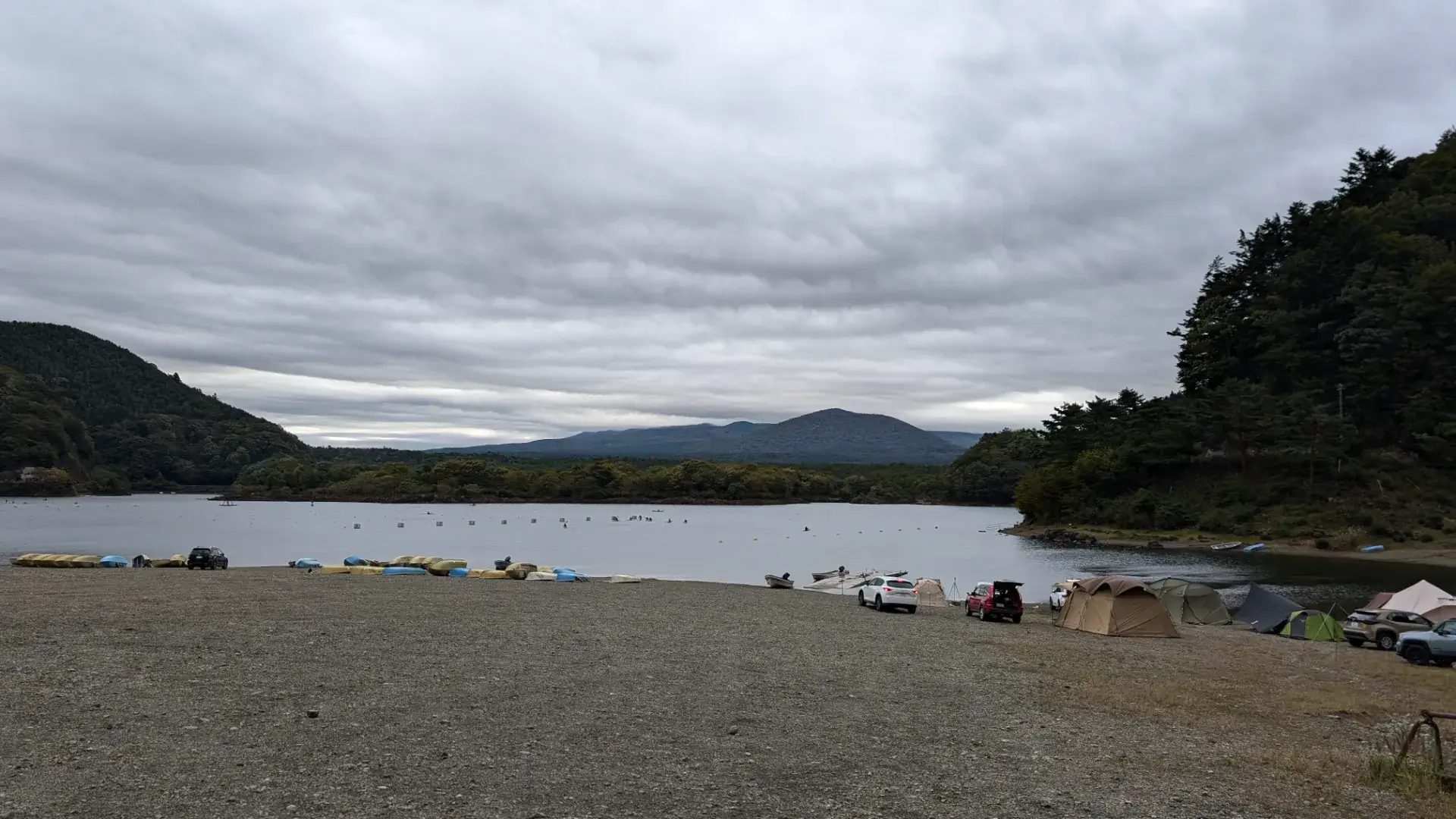 山梨県の精進湖(しょうじこ)にある他手合浜(たてごうはま)でのキャンプ風景。曇り空の下、湖畔に並ぶテントや車、ボート、そして遠くに広がる山々の景色。富士山を望む絶好のキャンプスポットとしての賑わい。