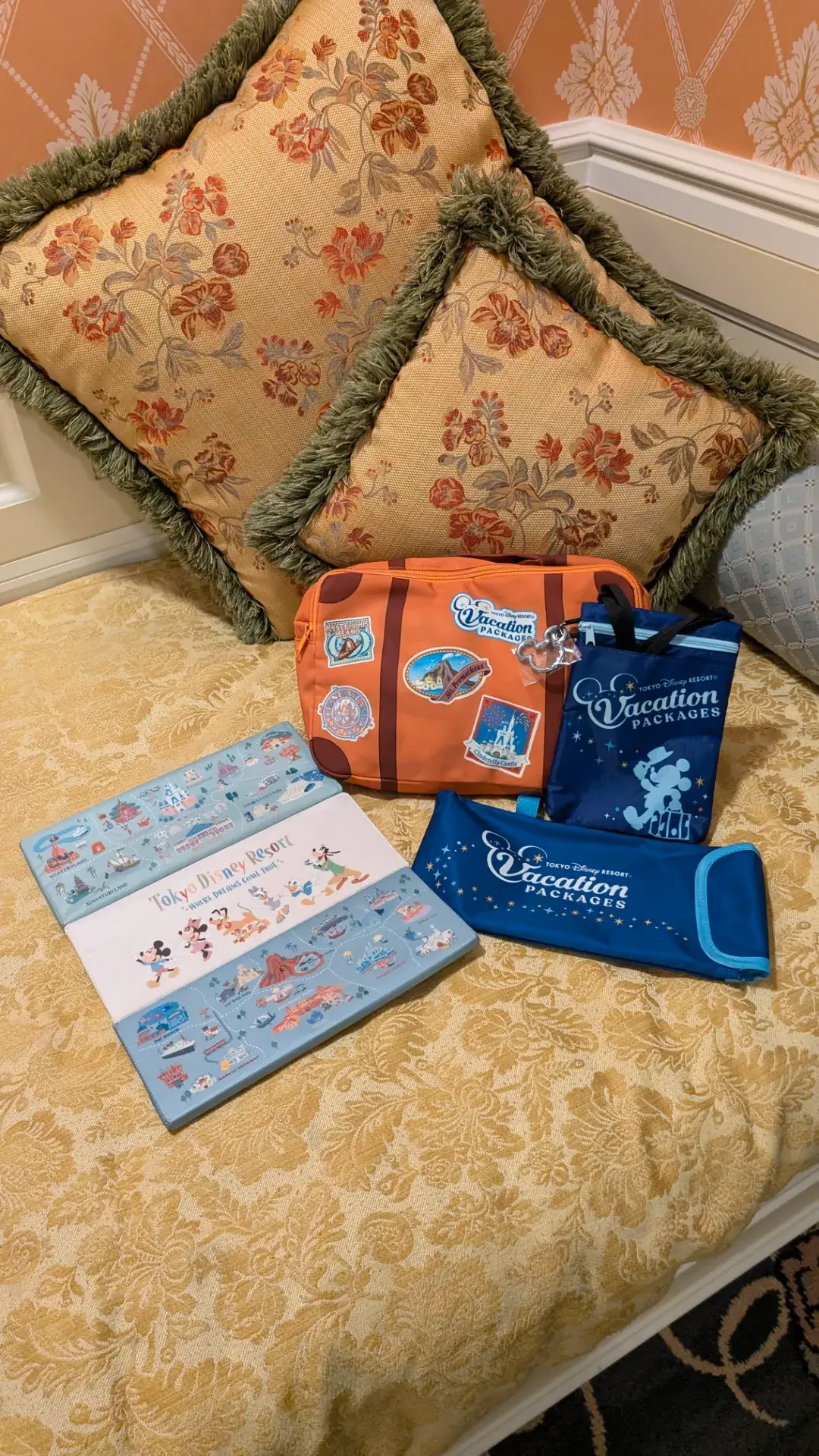 東京ディズニーランドホテルのアルコーヴベッドの上に並べられた、バケーションパッケージ限定のオリジナルグッズ。ミッキーのシルエットが描かれたネイビーのショルダーバッグやステーショナリーセット、様々なパークモチーフのステッカーがデザインされたオレンジのポーチなどが、華やかなクッションと共に配置されている。