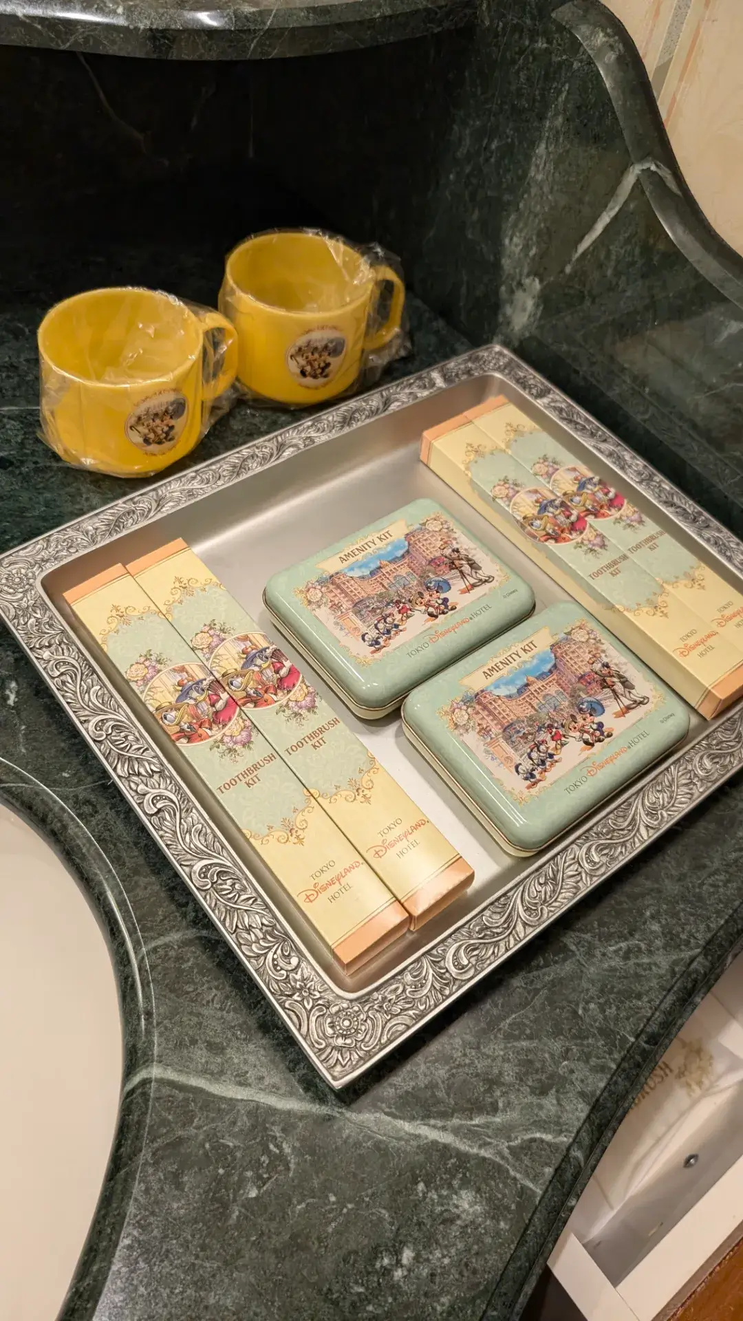 東京ディズニーランドホテルの客室アメニティ。ミッキー＆フレンズとホテル外観が描かれた可愛らしい缶入りアメニティキット、歯ブラシセット、そしてホテルのロゴが入った黄色いプラスチックコップがシルバーのトレイに美しく並べられている。