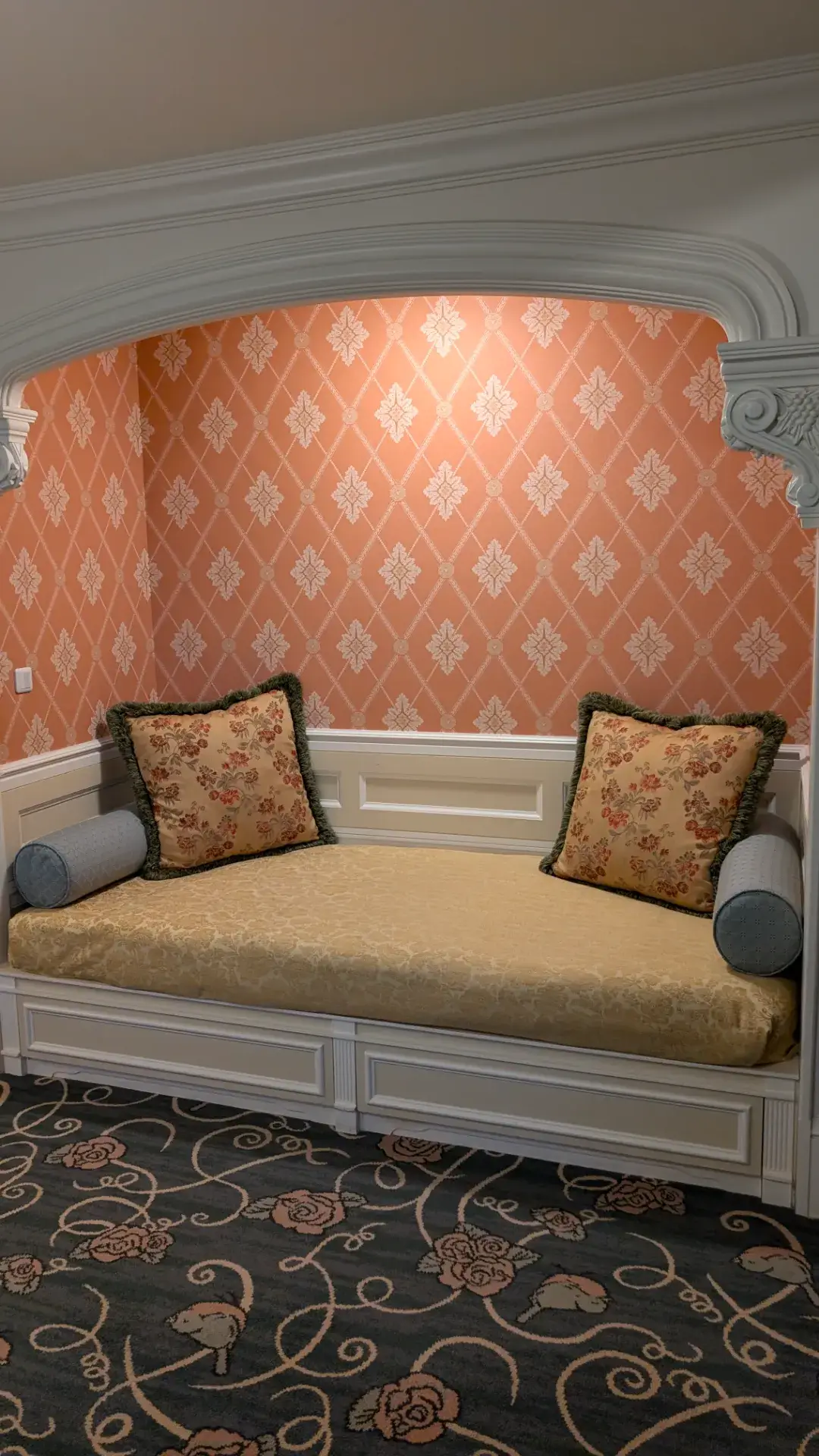 東京ディズニーランドホテルの客室インテリア。バラの模様が施されたブルーの絨毯に、クラシックなデザインのソファとアームチェアが配置されたリビングスペース。優雅な壁紙、アンティーク調のテレビ台やサイドテーブル、柔らかな光のランプが高級感あふれる空間を演出している。