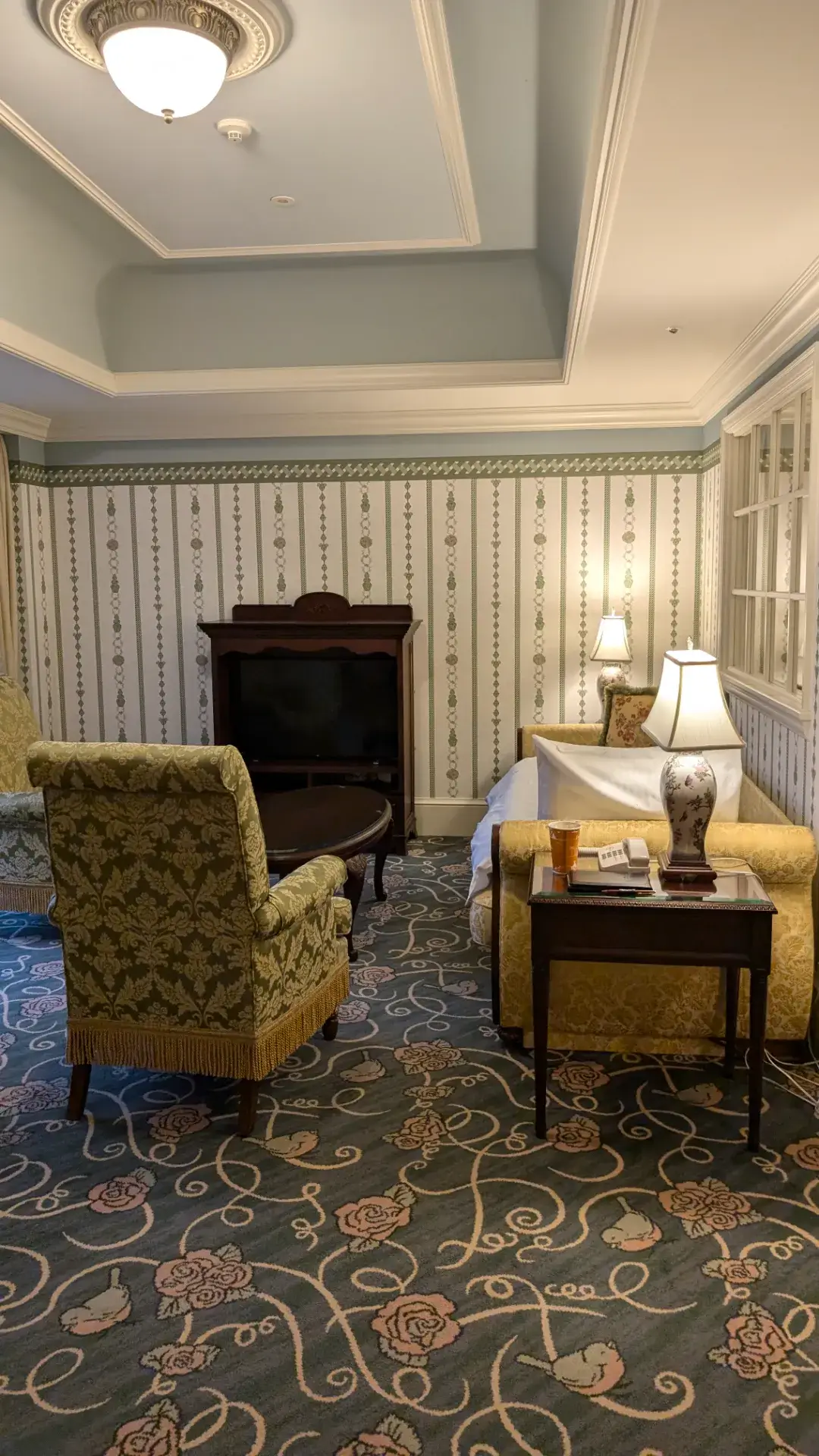 東京ディズニーランドホテルの客室インテリア。バラの模様が施されたブルーの絨毯に、クラシックなデザインのソファとアームチェアが配置されたリビングスペース。優雅な壁紙、アンティーク調のテレビ台やサイドテーブル、柔らかな光のランプが高級感あふれる空間を演出している。