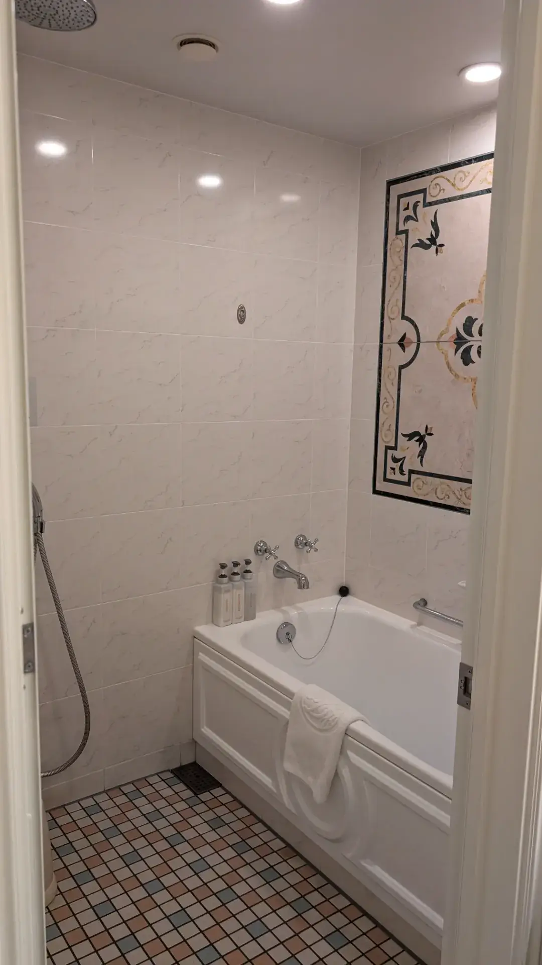 東京ディズニーランドホテルのバスルーム。大理石調の白い壁に施された美しいモザイクアート、広々とした白いバスタブ、そしてカラフルな小石のような床タイルが特徴的な、エレガントで清潔感あふれる空間。