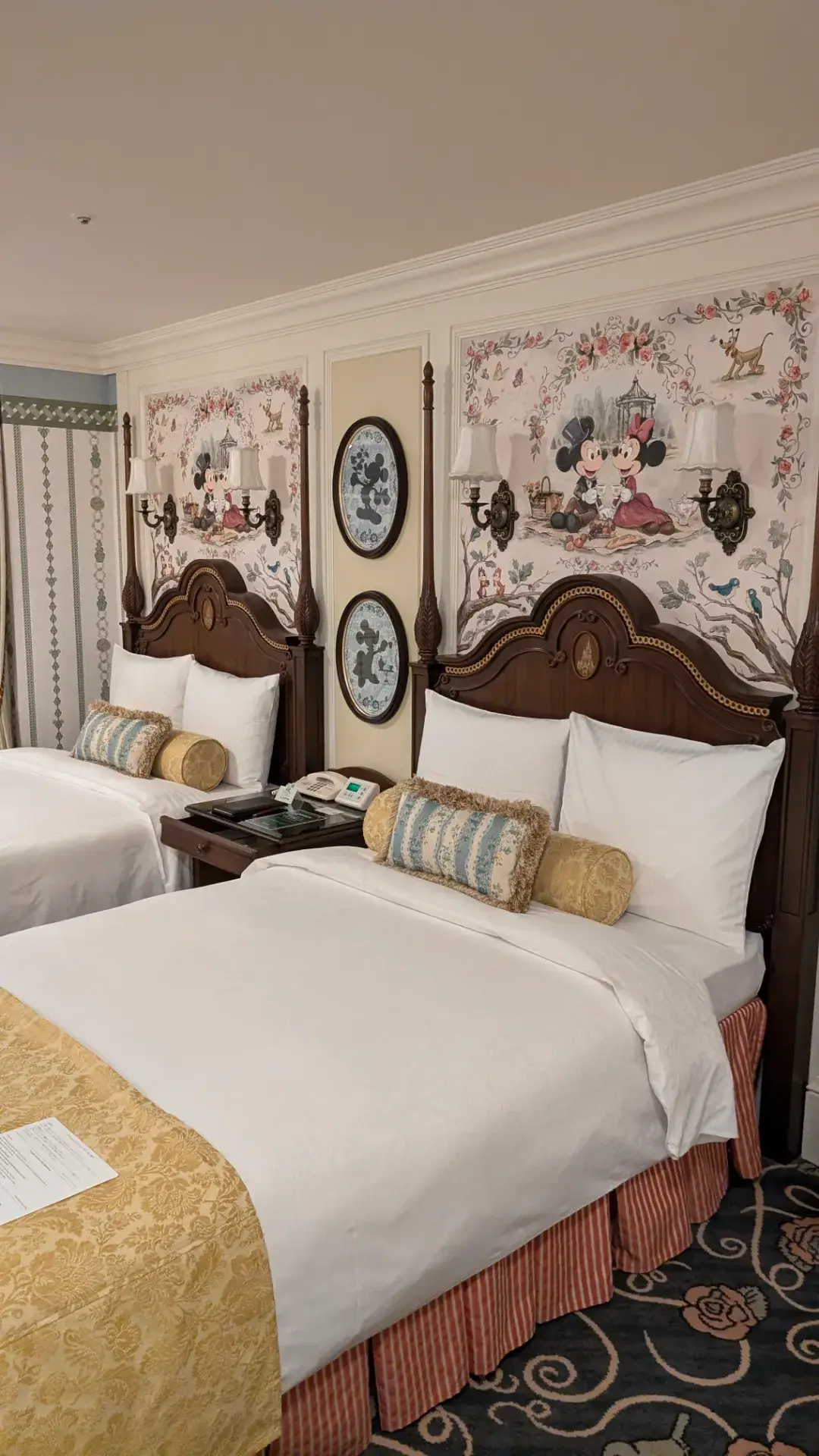 東京ディズニーランドホテルの客室。ヘッドボードの上の壁面には、ピクニックを楽しむミッキーとミニーのクラシックな壁画が描かれている。エレガントな木製のツインベッドには、上品な刺繍のクッションとフットスローが整えられ、魔法の国にふさわしい夢のような空間。