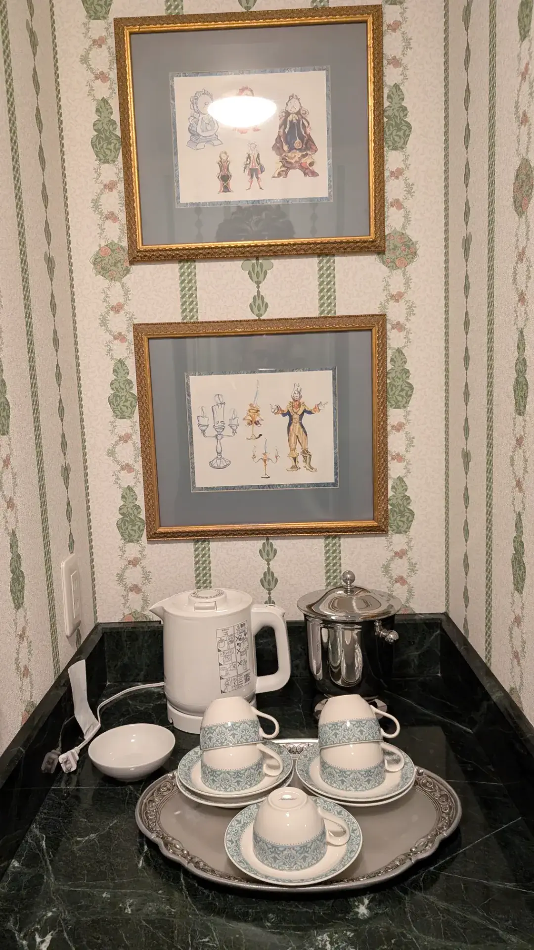 東京ディズニーランドホテルの客室にあるティーカウンター。壁には映画『美女と野獣』のキャラクター、コグスワースやルミエールたちの設定画が飾られ、大理石のカウンターには電気ケトル、アイスペール、そしてエレガントな模様のティーカップセットが用意されている。