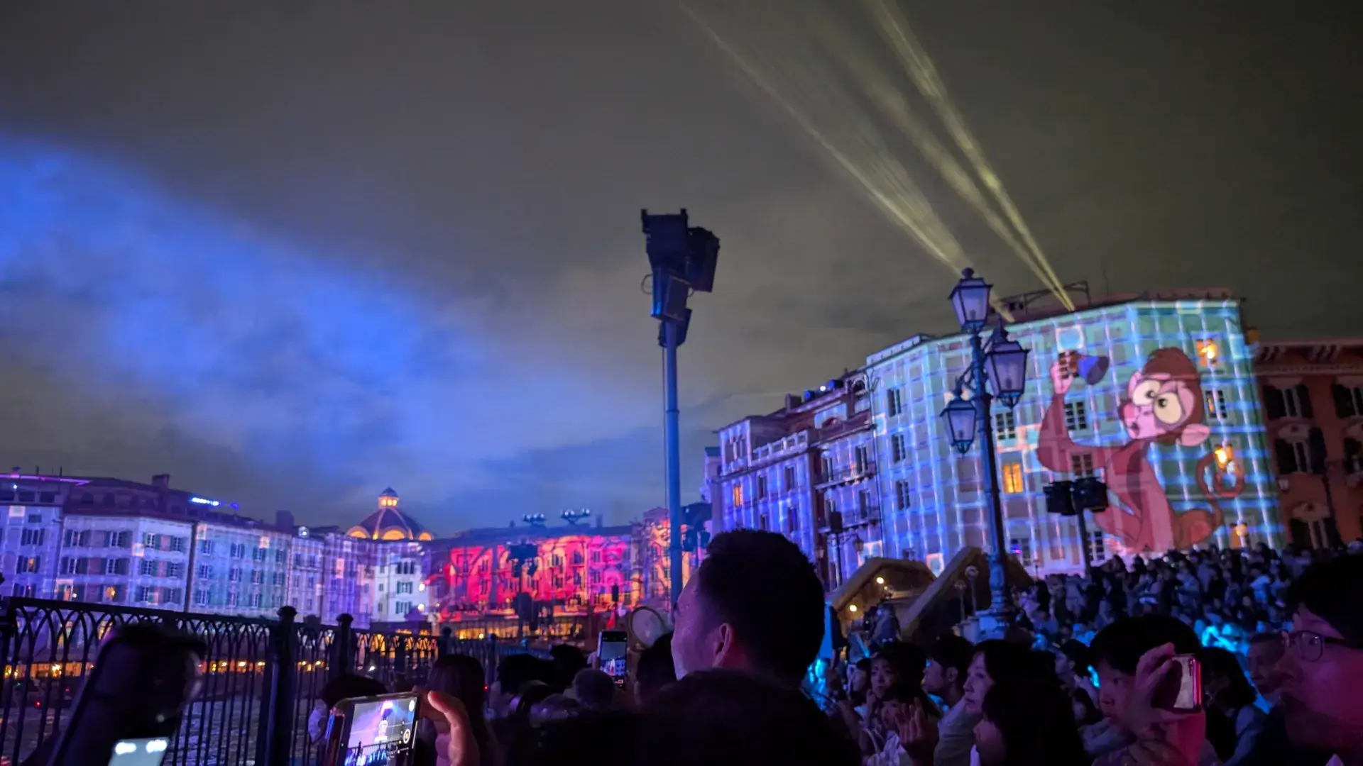 夜の東京ディズニーシー。ホテル・ミラコスタの壁面に、映画『アラジン』に登場する猿のアブーが大きく映し出されているプロジェクションマッピング。サーチライトが夜空を照らし、大勢の観客がスマートフォンでその壮大な光のショーを記録している。