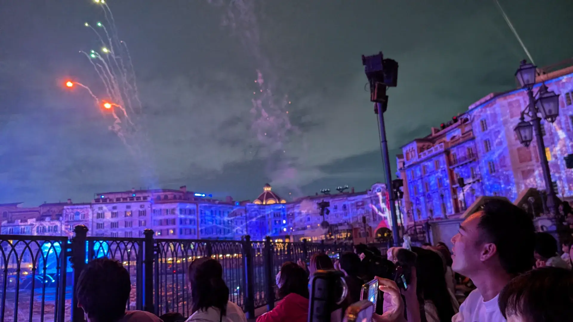 東京ディズニーシーのナイトショー「ビリーヴ！」のクライマックス。中央のタワーから高く打ち上がる無数のパイロ（花火）が夜空を白く照らし、ハーバーのフェンス越しにショーを鑑賞する人々のシルエットがその輝きを際立たせている。