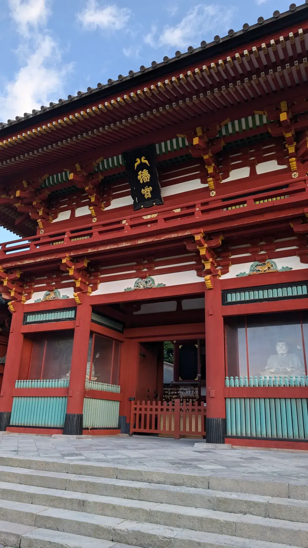 神奈川県鎌倉市の鶴岡八幡宮(つるおかはちまんぐう)にある鮮やかな朱塗りの楼門。青空を背景に、歴史を感じさせる緻密な彫刻と「八幡宮」と書かれた黒い扁額(へんがく)、門の左右に鎮座する随身像が写っている。