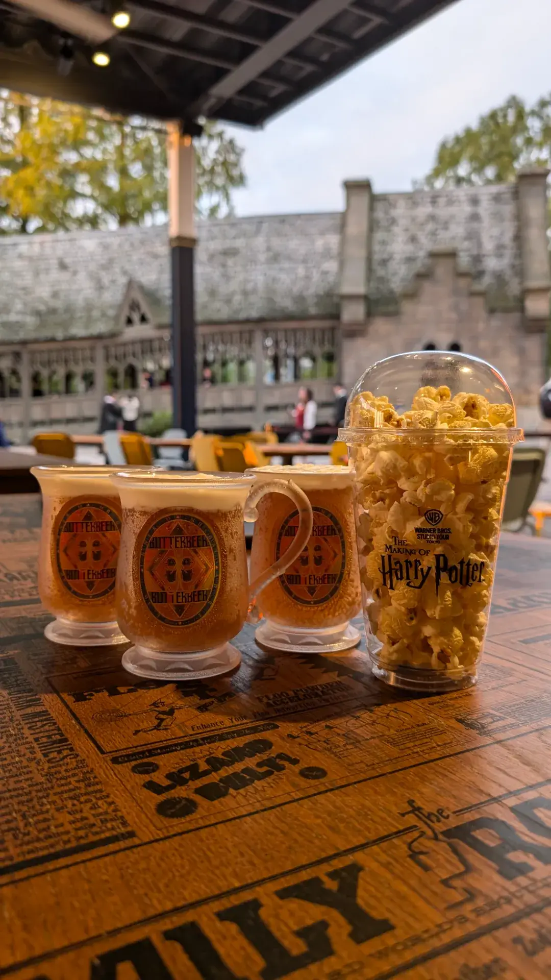 ワーナー ブラザース スタジオツアー東京にて、魔法界の人気飲み物「バタービール」が3つと、ハリー・ポッターのロゴ入りカップに入ったポップコーンが並んでいる様子。テーブルには映画の世界観を再現した英字新聞風のプリントが施されている。