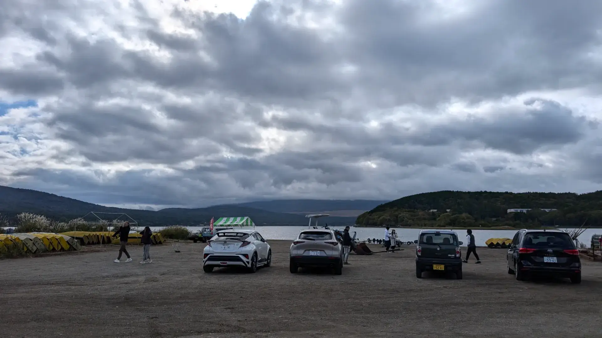 山梨県山中湖村の「平野の浜」の様子。曇り空の下、湖畔の砂浜に駐車された車や、穏やかな湖を背景に散策を楽しむ人々の姿が見られる。富士山ビュースポットとして知られる開放的なロケーション。