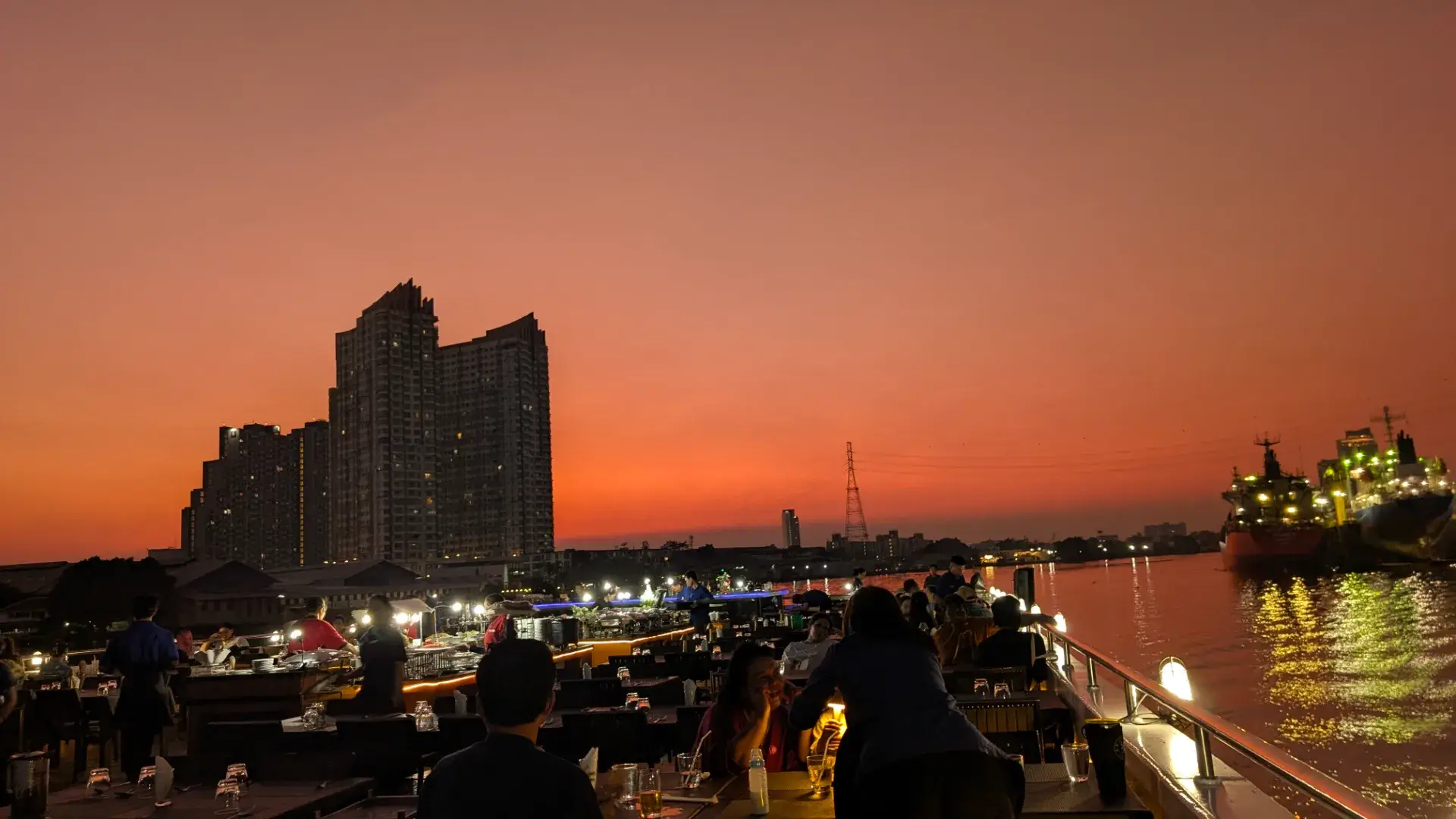 バンコクのチャオプラヤー川、ディナークルーズ船のデッキからの眺め。地平線が鮮やかなオレンジ色に染まるマジックアワーの中、シルエットとなった高層ビル群と、ライトアップされて水面に光を落とす大きな貨物船が作り出すドラマチックな夜景。