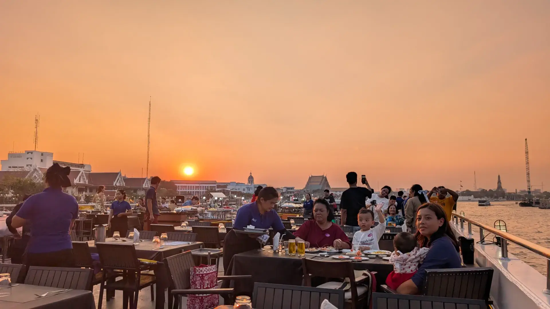 タイ・バンコク、チャオプラヤー川のディナークルーズ船のデッキ。オレンジ色に染まる美しい夕日を背景に、多くのゲストがテーブルを囲み、スタッフが忙しく立ち働く活気ある夕暮れ時の様子。