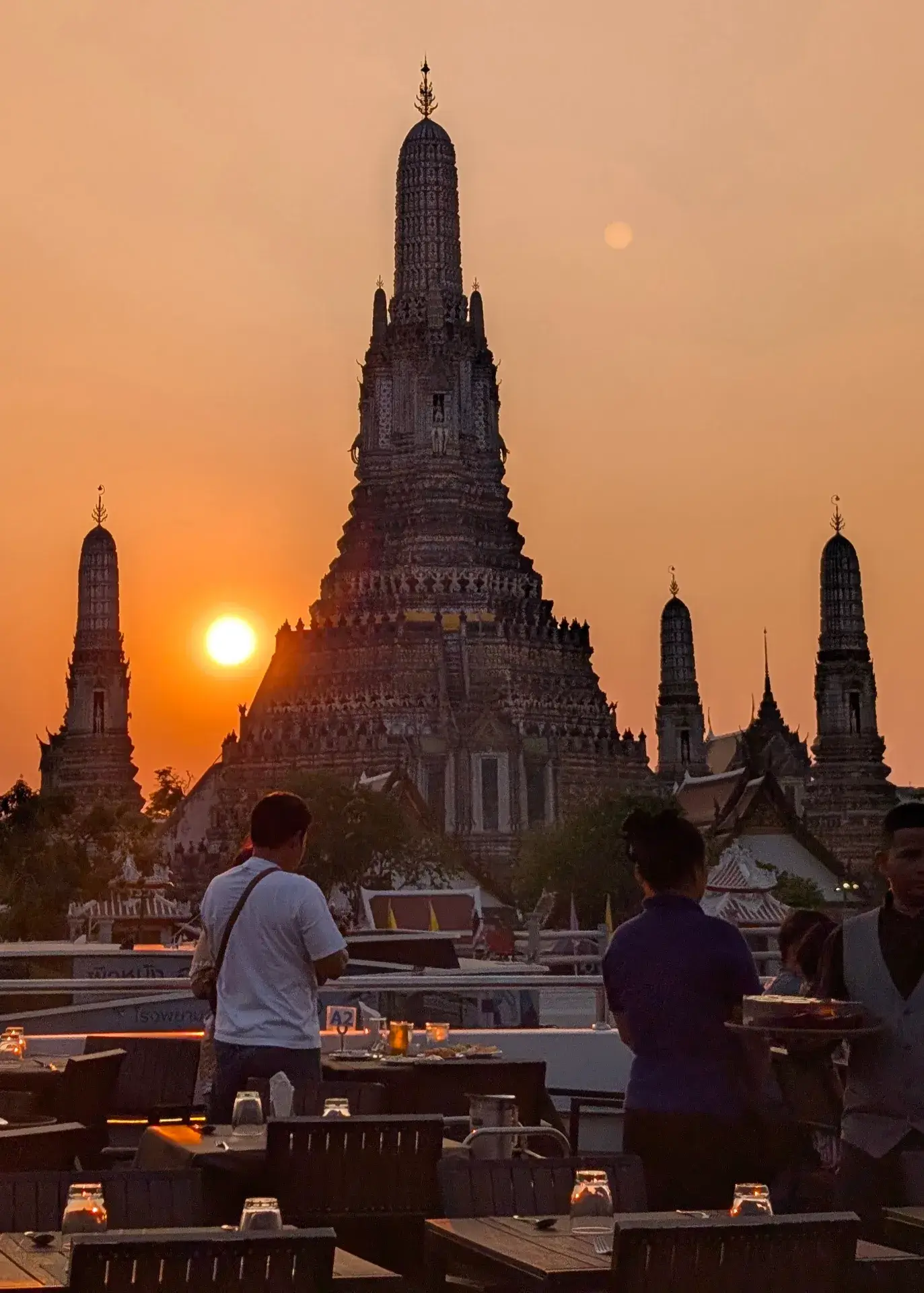 バンコクのチャオプラヤー川を航行するディナークルーズ船から見た、夕暮れ時のワット・アルン。巨大な仏塔（大プラーン）のシルエットがオレンジ色の空に映え、沈みゆく太陽が寺院の横で輝く幻想的な風景。手前にはディナーを楽しむ船上のゲストの姿が見える。