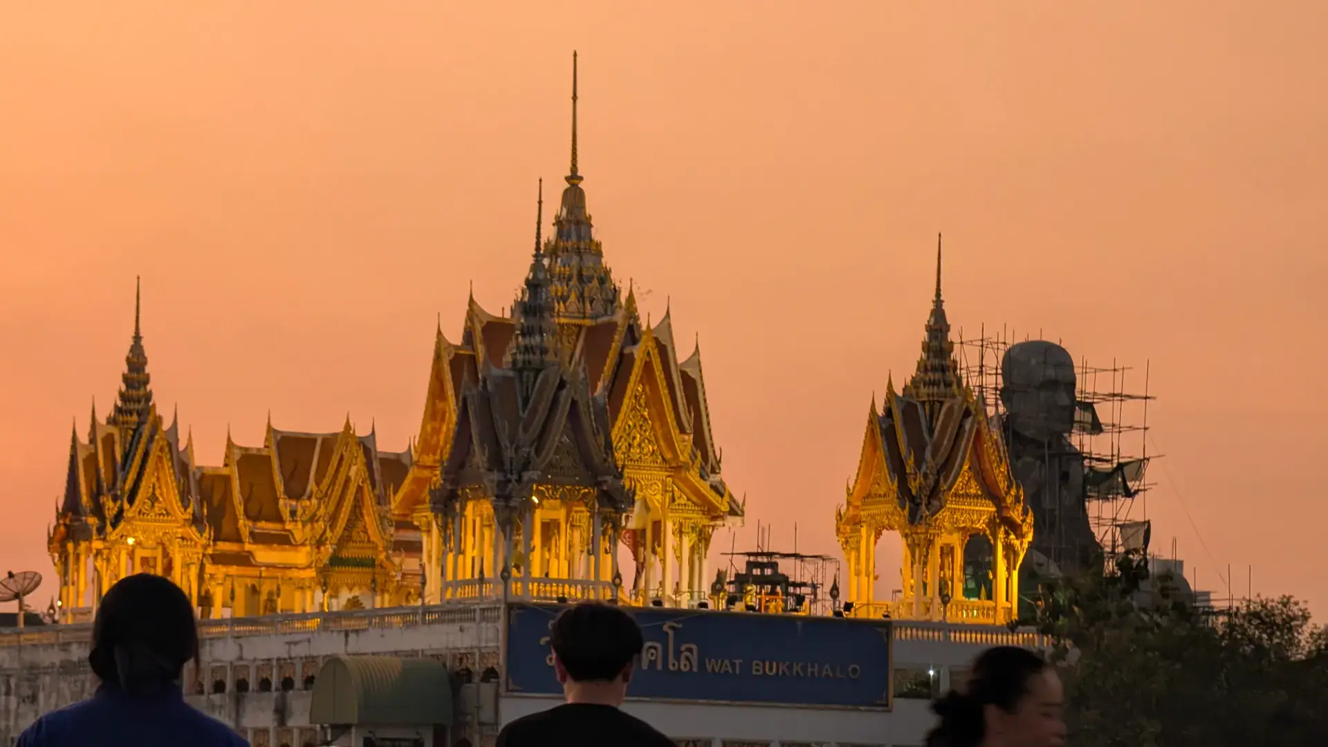 タイ・バンコク。チャオプラヤー川のディナークルーズから望む、夕日に照らされたワット・ブックカロの美しい寺院。伝統的なタイ様式の鋭い屋根が黄金色に輝き、右奥には建設中の巨大な大仏の姿が見える、ドラマチックな夕暮れの風景。