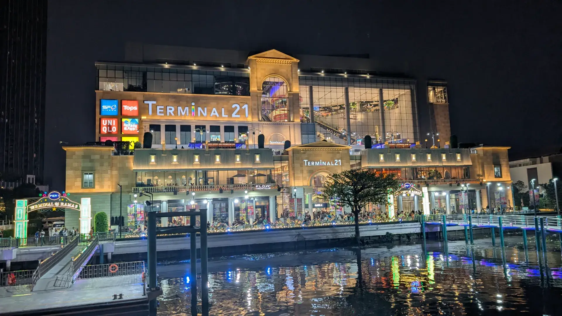タイ・バンコクのチャオプラヤー川沿いに建つショッピングモール「TERMINAL 21 RAMA 3」の豪華な夜景。ライトアップされた巨大な建物、UNIQLOやTopsなどの看板、そして川面に反射する色鮮やかな光のネオンが美しい、現代的なリバーサイドの風景。