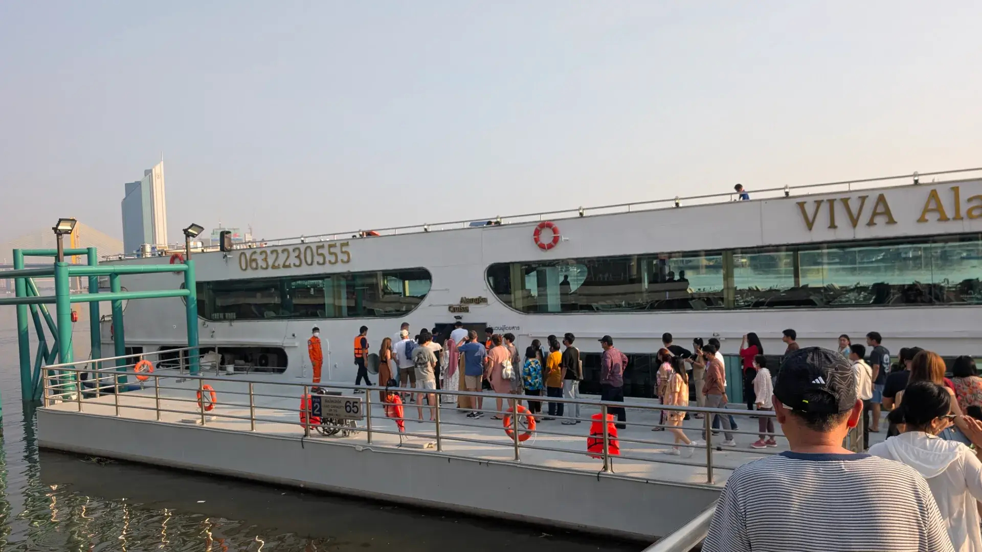 タイ・バンコクのチャオプラヤー川沿い。夕暮れ時、多くの観光客が大きな白いクルーズ船「VIVA Alangka Cruise」に乗り込もうと桟橋に並んでいる様子。背景には近代的なビルや橋が見える、賑やかなリバーサイドの風景。