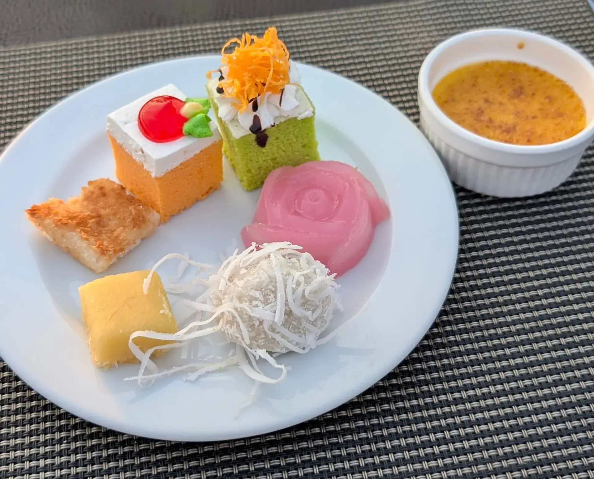 バンコクのディナークルーズで提供されたデザートプレート。ピンク色のバラの形をしたカノムチャン（ういろう風菓子）、ココナッツをまぶしたルクチュップ、色鮮やかなミニケーキなどのタイ伝統菓子と、横に添えられたカスタードプリン（カノムモーケン）。