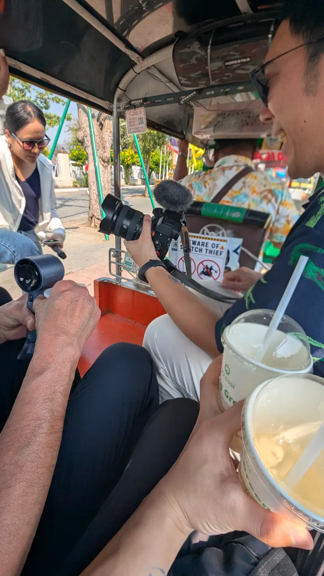 バンコク観光の醍醐味であるトゥクトゥクに乗り、風を切って街を移動するエキサイティングな瞬間。手元には「DOI KHAM」のドリンク、そして首からは旅の決定的瞬間を逃さないためのミラーレスカメラが下げられています。車内には「ひったくり注意」のステッカーも見え、活気ある街の熱気を感じながら次の目的地へと向かう、旅の躍動感が詰まった一枚です。