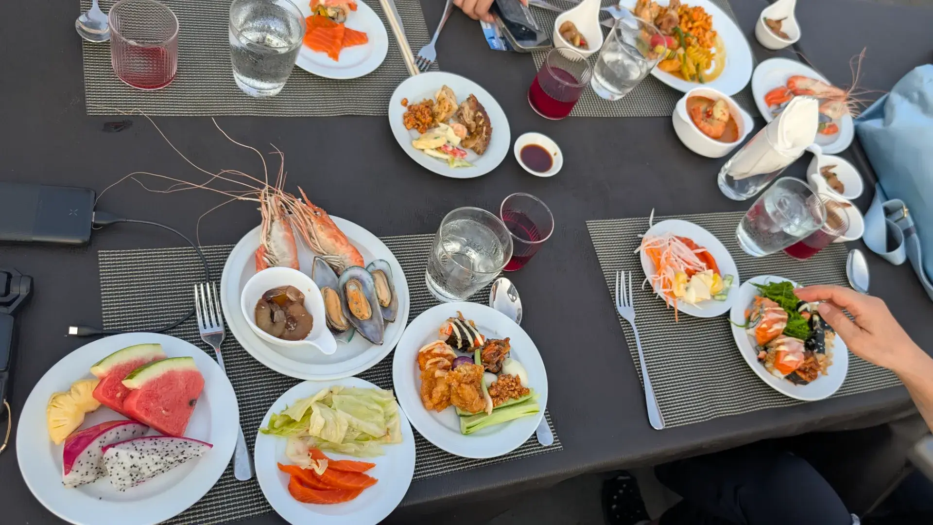 バンコクのチャオプラヤー川クルーズ船上でのディナー風景。テーブルには、大きな海老（エビ）やムール貝の盛り合わせ、サーモンの刺身、ドラゴンフルーツやスイカなどの新鮮なフルーツ、さらに寿司や温菜が並ぶ豪華なビュッフェ料理の数々。