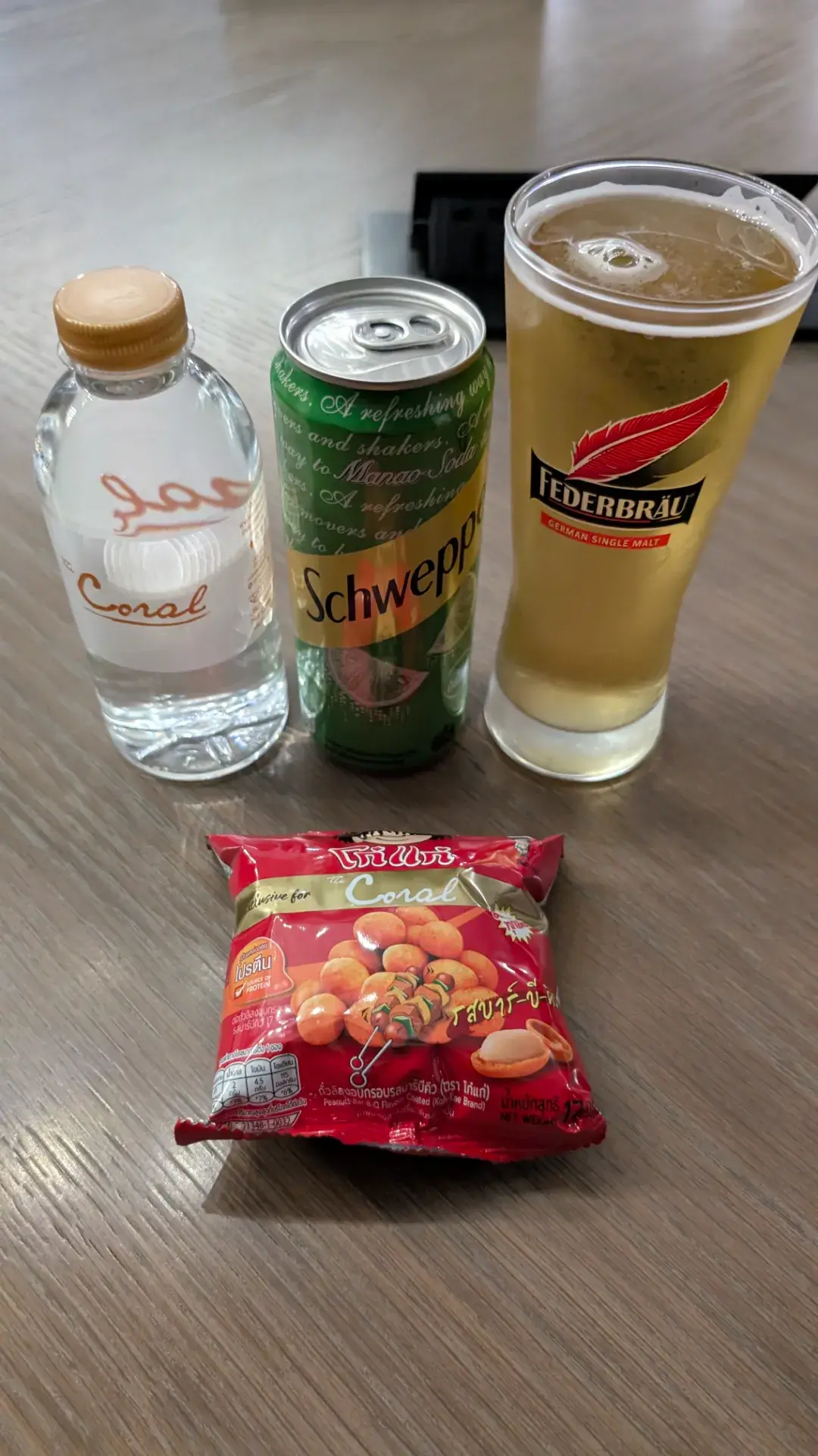 搭乗前のひととき、ドンムアン空港のラウンジで楽しむリフレッシュメント。ラウンジオリジナルのミネラルウォーター、シュウェップスのマナオ（タイライム）ソーダ、そして「Federbräu（フェーダーブロイ）」の生ビールが並びます。手前には、ラウンジ限定パッケージの「Koh-Kae（コーゲー）」の豆菓子も添えられ、タイでの美食の思い出を締めくくる、穏やかで贅沢な待ち時間の一コマです。