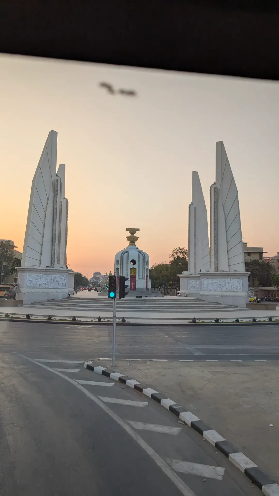 バンコクのラチャダムヌン通りに建つ「民主記念塔」を、観光バスの車内から捉えた風景。夕日に照らされた4つの巨大な翼状の塔と、中央の憲法を安置した台座が、オレンジ色の空に美しく映えています。バスツアーのルート上でも特に存在感を放つランドマークであり、タイの近代史を感じさせるとともに、刻一刻と表情を変えるバンコクの夕景を堪能できるハイライトシーンです。