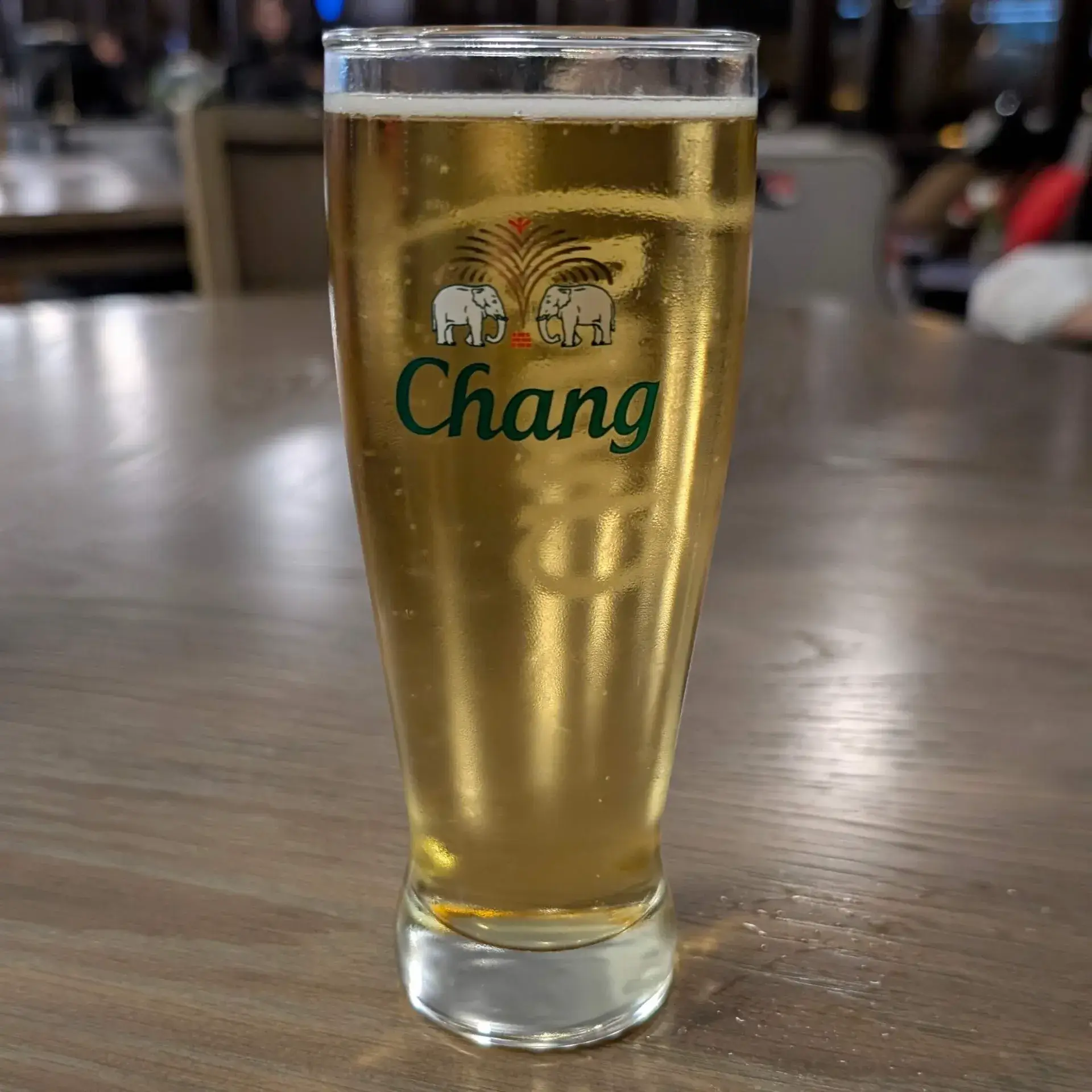 ドンムアン空港のコーラル・ラウンジにて、最後の一杯として楽しむタイの国民的ビール「チャーン（Chang）」。トレードマークである2頭の白い象が描かれたグラスに、黄金色のビールが美しく輝いています。スコータイの遺跡からバンコクの賑やかな夜まで、数々の思い出を語り合いながら味わう喉越しの良い一杯は、タイ旅行のフィナーレを最高な形で締めくくってくれます。