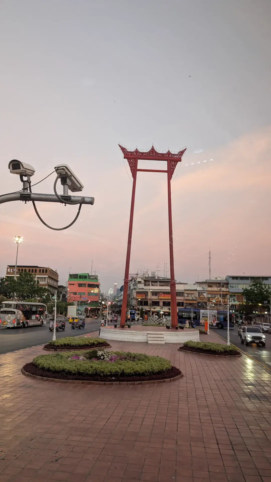 「タイ・バス・フードツアー」の車窓から眺める、バンコクのランドマーク「ジャイアント・スイング（サオ・チン・チャー）」。かつてバラモン教の儀式に使われていた高さ21メートルを超える巨大な赤い柱が、夕暮れ時の幻想的な空に凛として立っています。周囲の歴史的な街並みと、近代的な監視カメラや走行するバスが交差するこの光景は、古き良き伝統と現代が共存するバンコクの奥深さを象徴する、旅のフィナーレを飾るにふさわしい一枚です。