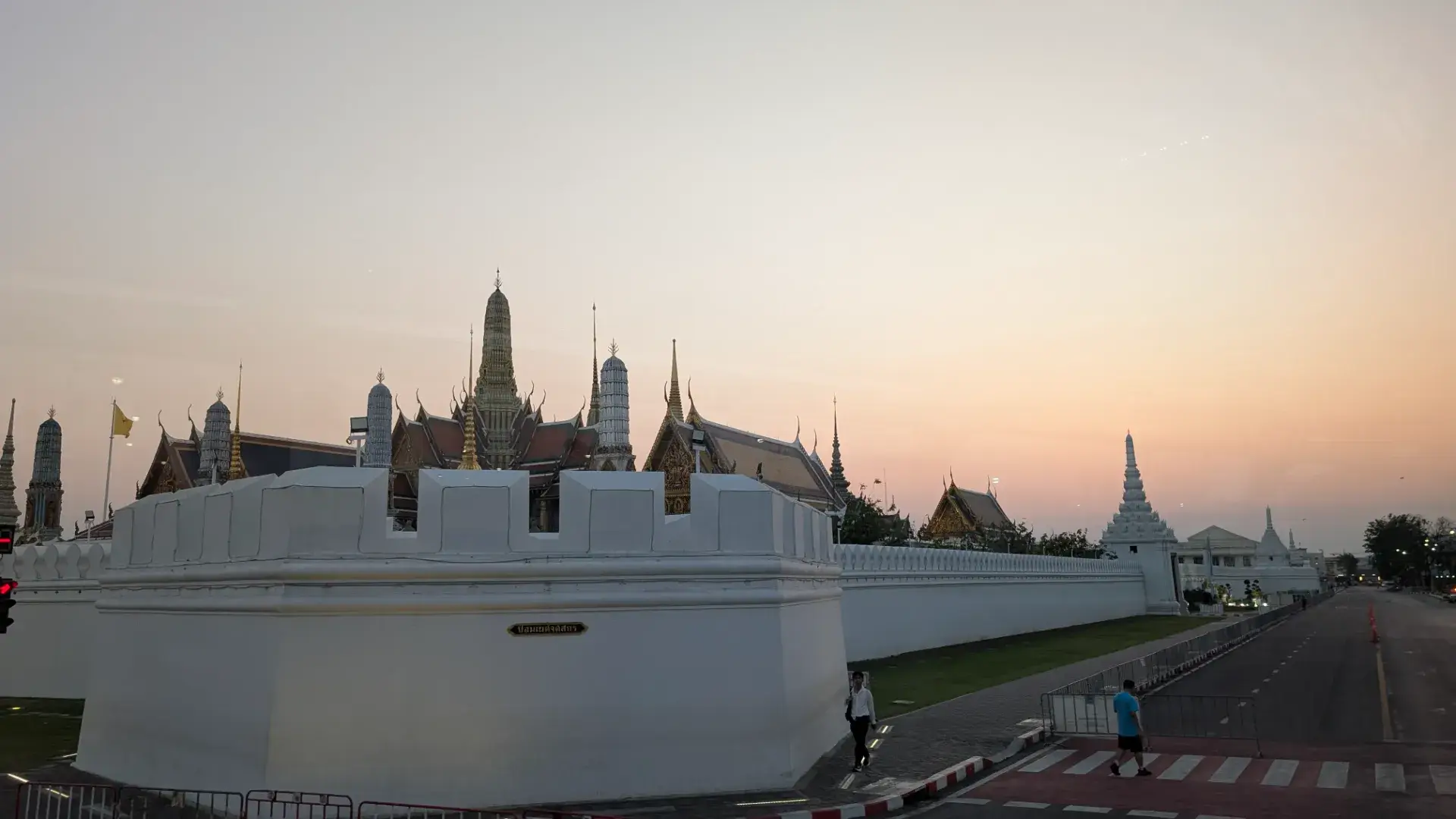 「タイ・バス・フードツアー」の車窓から望む、黄昏時のワット・プラケオ（エメラルド寺院）。白い城壁越しに、タイ伝統建築の粋を集めた黄金の仏塔や重層的な屋根が、淡いピンク色の空に溶け込むようにそびえ立っています。日中の喧騒が去り、静寂とともにライトアップが灯り始めるこの瞬間の王宮は、バスツアーの中でも息を呑むほど幻想的な美しさを放つ絶景ポイントです。