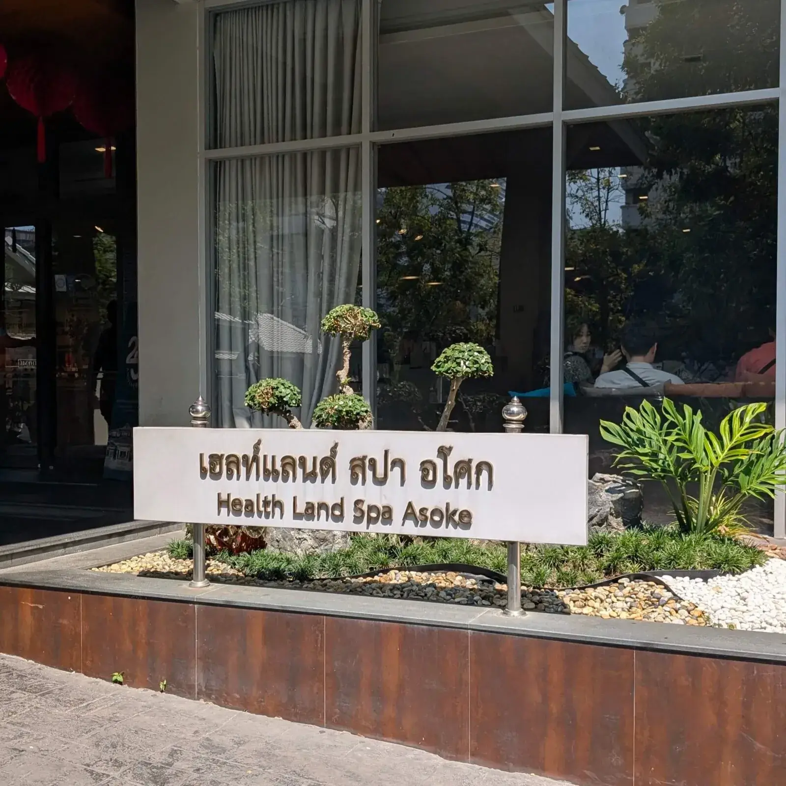 バンコクのアソークエリアに位置する、有名スパ「ヘルスランド（Health Land）」の看板。手入れの行き届いた植栽とともに、「Health Land Spa Asoke」とタイ語・英語で記された白い看板が、訪れる人を清潔感のある落ち着いた空間へと迎え入れます。高品質なマッサージを手頃な価格で受けられることで、観光客から地元の人々まで幅広く愛されているリラクゼーションの名所です。