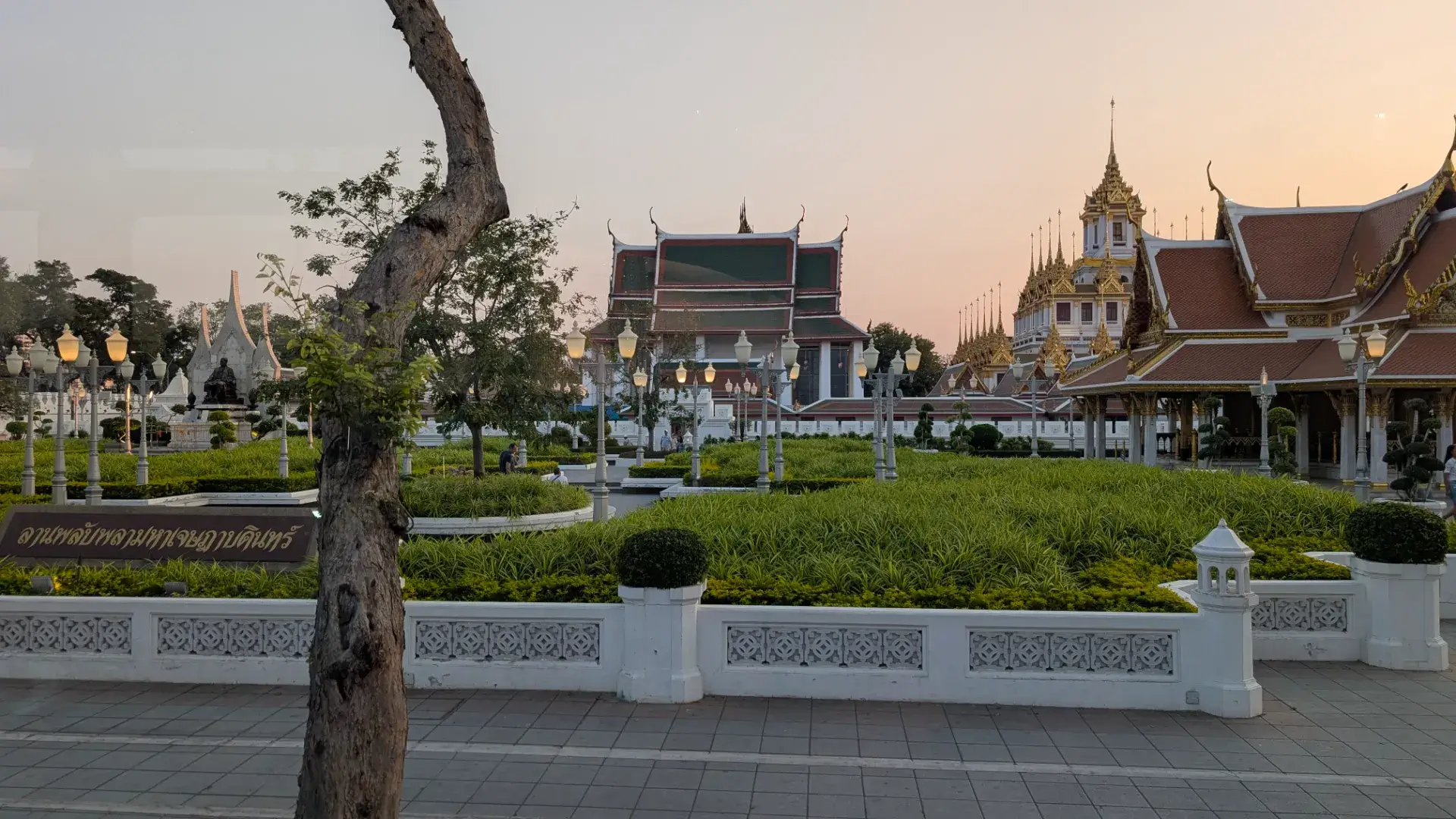 「タイ・バス・フードツアー」の走行中に現れる、別名「鉄の城」とも呼ばれる仏塔「ロハ・プラサート」。37本の尖塔が空に伸びる独特の建築様式が、夕暮れの柔らかな光に包まれ、幻想的なシルエットを浮かび上がらせています。美しく整えられた庭園と、伝統的なタイ寺院の屋根が織りなすこの風景は、車内での優雅な食事にさらなる彩りを添える、ツアー屈指の絶景ポイントです。
