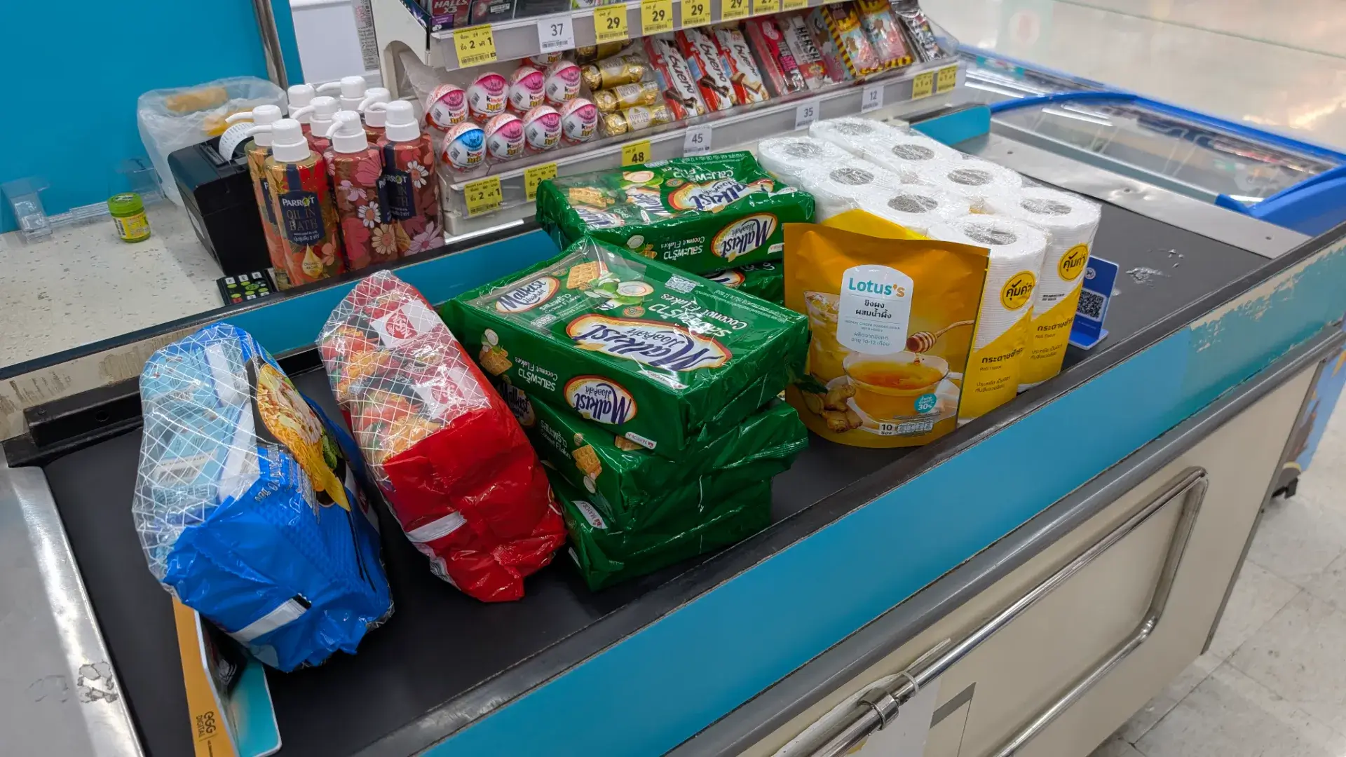 タイの大手スーパーマーケット「Lotus's（ロータス）」のレジカウンターに並ぶ購入品。タイで定番のクラッカー「Malkist（マルキスト）」の大袋やトイレットペーパー、Lotus'sプライベートブランドのハチミツ生姜茶など、現地での生活感が伝わる品々が並んでいます。旅の締めくくりにお土産や日用品を賢く揃える、タイ旅行のリアルなワンシーンです。