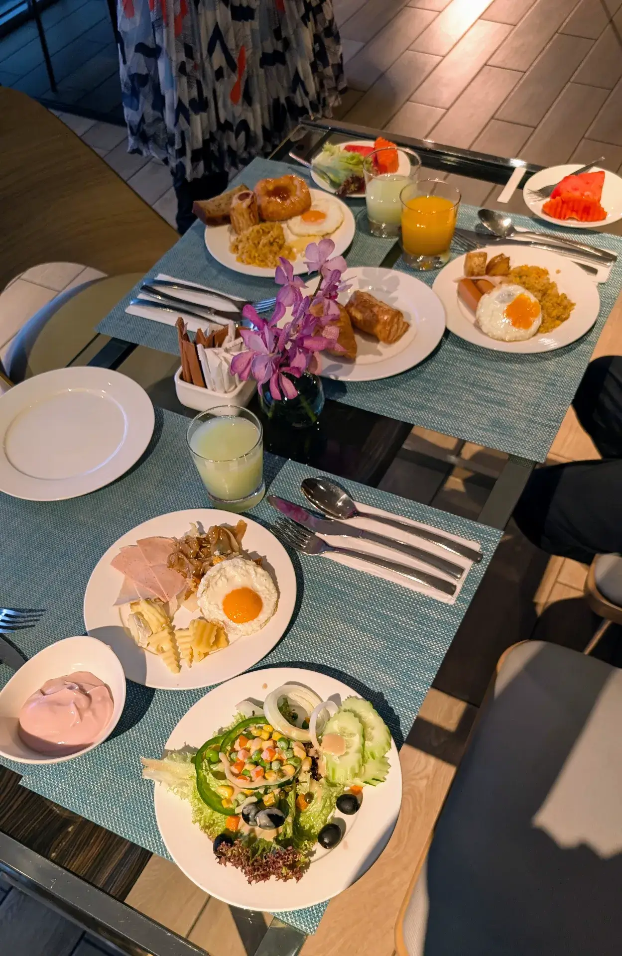「モデナ バイ フレイザー バンコク」で楽しむ贅沢な朝食。テーブルには、目玉焼きを添えたタイ風の炒め物やソーセージ、焼きたてのデニッシュ、新鮮な生野菜サラダ、そしてスイカやオレンジジュースなどのトロピカルなメニューが並びます。紫色の蘭の花が添えられた、清潔感と華やかさのある朝のひとときです。
