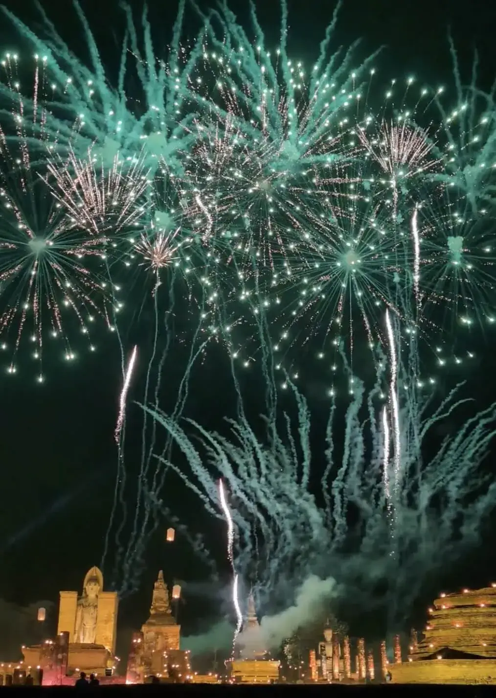 夜のスコータイ歴史公園。ライトアップされた仏像と仏塔の真上に、鮮やかなエメラルドグリーンの花火が大きく打ち上がっている。無数の光の筋が夜空を埋め尽くし、幻想的な緑の煙がたなびく祝祭の瞬間。