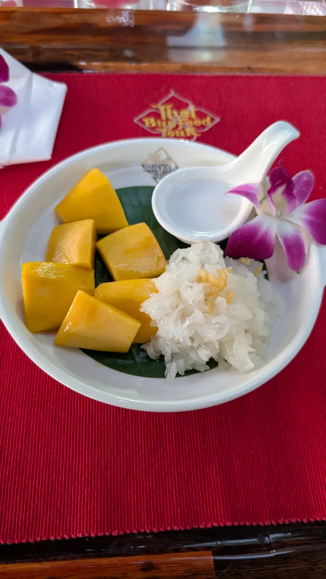 「タイ・バス・フードツアー」のコースを締めくくる、タイの代表的なデザート「カオニャオ・マムアン」。完熟した黄金色のマンゴーに、ココナッツミルクで炊き上げた甘いもち米が添えられています。もち米の上にはカリカリとした緑豆のトッピングが施され、食感のアクセントに。紫色の蘭の花が美しく飾られた一皿は、車窓に流れるバンコクの夜景とともに味わう、至福の旅のエンディングを演出してくれます。