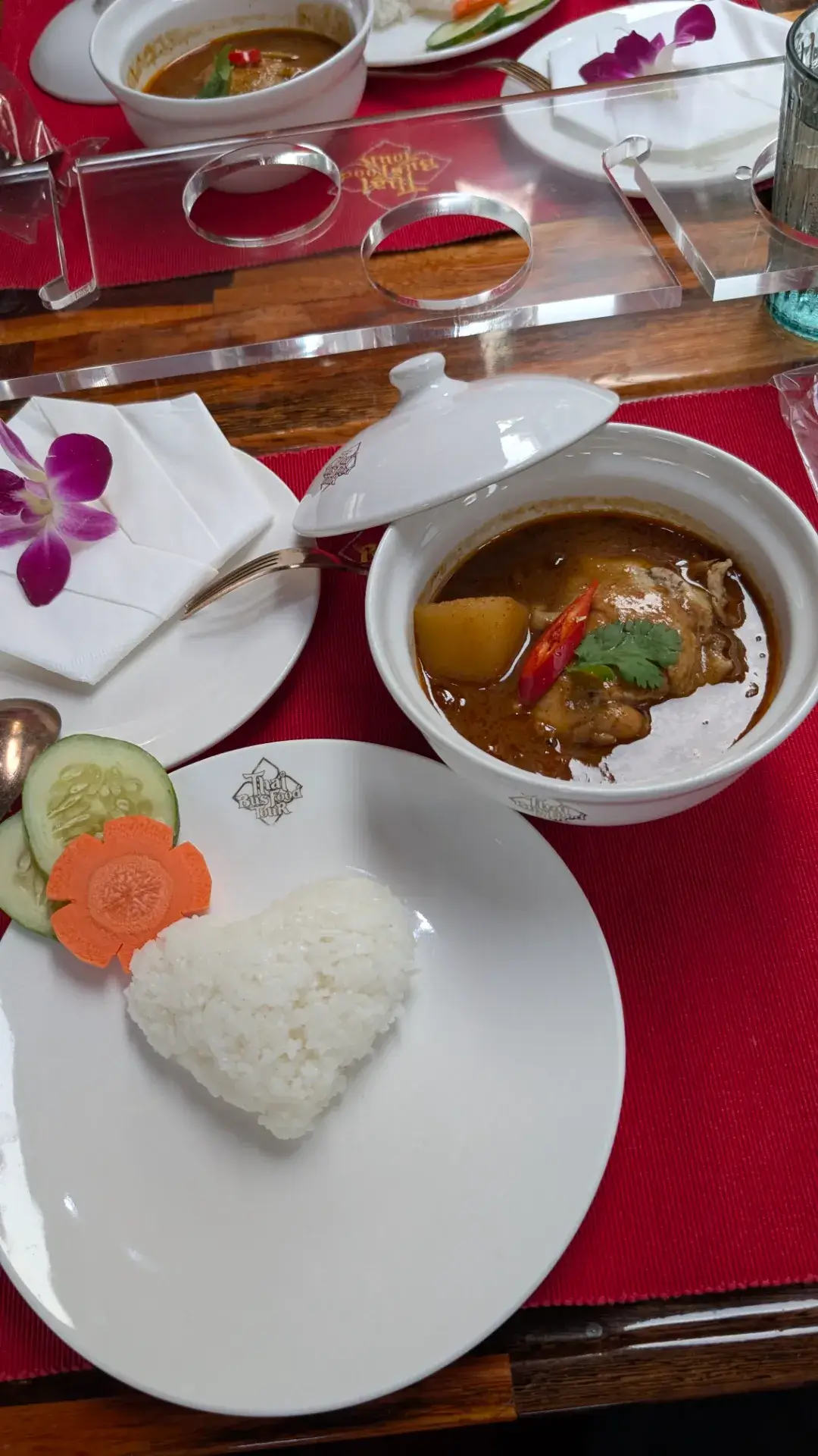 「タイ・バス・フードツアー」のコースを彩るメイン料理、鶏肉のマッサマンカレー。ココナッツミルクの甘みとスパイスの深いコクが特徴のカレーには、ゴロゴロとした大きなジャガイモと柔らかな鶏肉が入り、食欲をそそります。添えられたライスは可愛らしいハート型に象られ、人参の花形の飾り切りとともに、バスツアーならではの細やかなホスピタリティを感じさせます。揺れる車内でも崩れない美しい盛り付けは、まさに旅の最高の思い出となる一皿です。