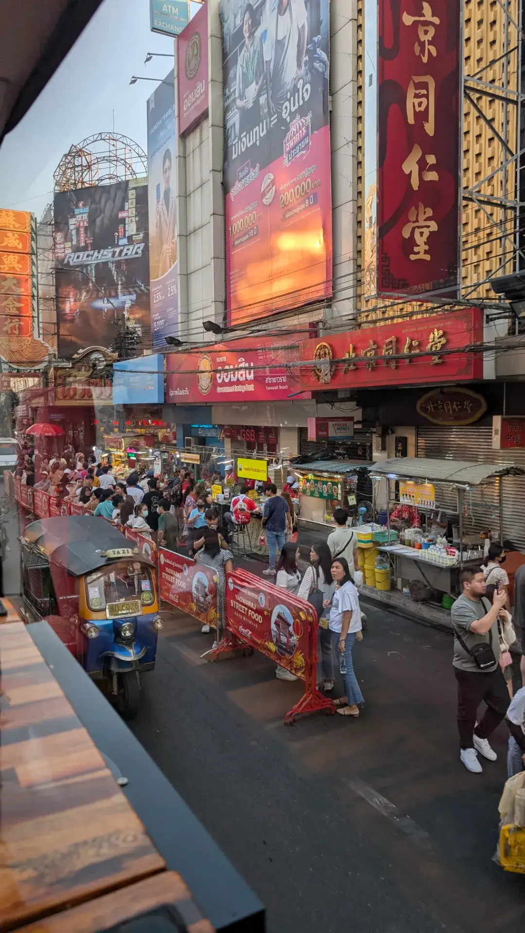 「タイ・バス・フードツアー」の2階席から見下ろした、バンコク・ヤワラート通りの活気ある光景。通りには名物のトゥクトゥクが走り、色鮮やかな看板が並ぶ中、多くの観光客や地元の人々がストリートフードを楽しんでいます。歩行者天国のように賑わう夕暮れ時の中華街を、涼しく快適な車内から贅沢に観光できる、バスツアーならではのダイナミックな視点の一枚です。