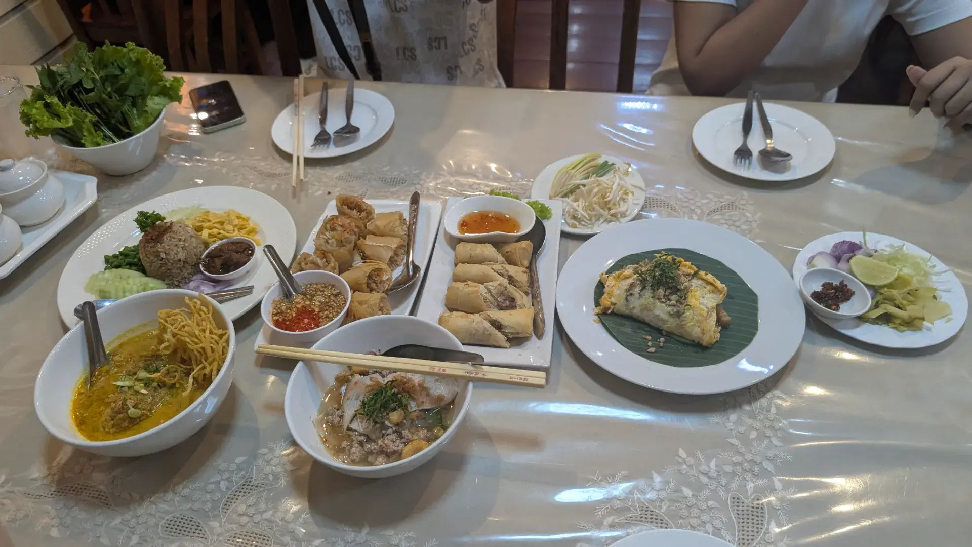 タイのレストランで提供された豪華なディナーの様子。北部の名物カレーうどん「カオソーイ」をはじめ、揚げ春巻き、タイ風オムレツのパッタイ、ハーブたっぷりのスープなど、色とりどりの本格タイ料理が並びます。家族や友人とシェアしながら、現地の味を存分に堪能できる賑やかな食卓の風景です。