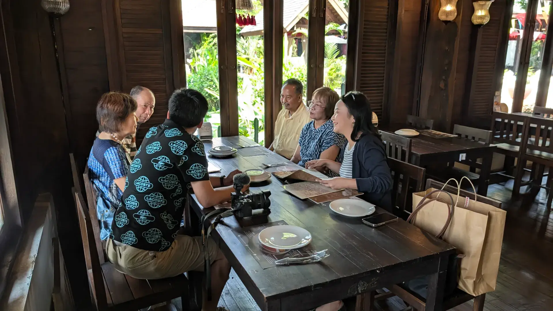 タイの伝統的な木造建築が美しいレストランでの会食風景。家族や友人とテーブルを囲み、メニューを広げながら談笑する和やかなランチタイムの様子です。落ち着いた内装と窓から差し込む光が、タイ旅行の豊かなひとときを演出しています。