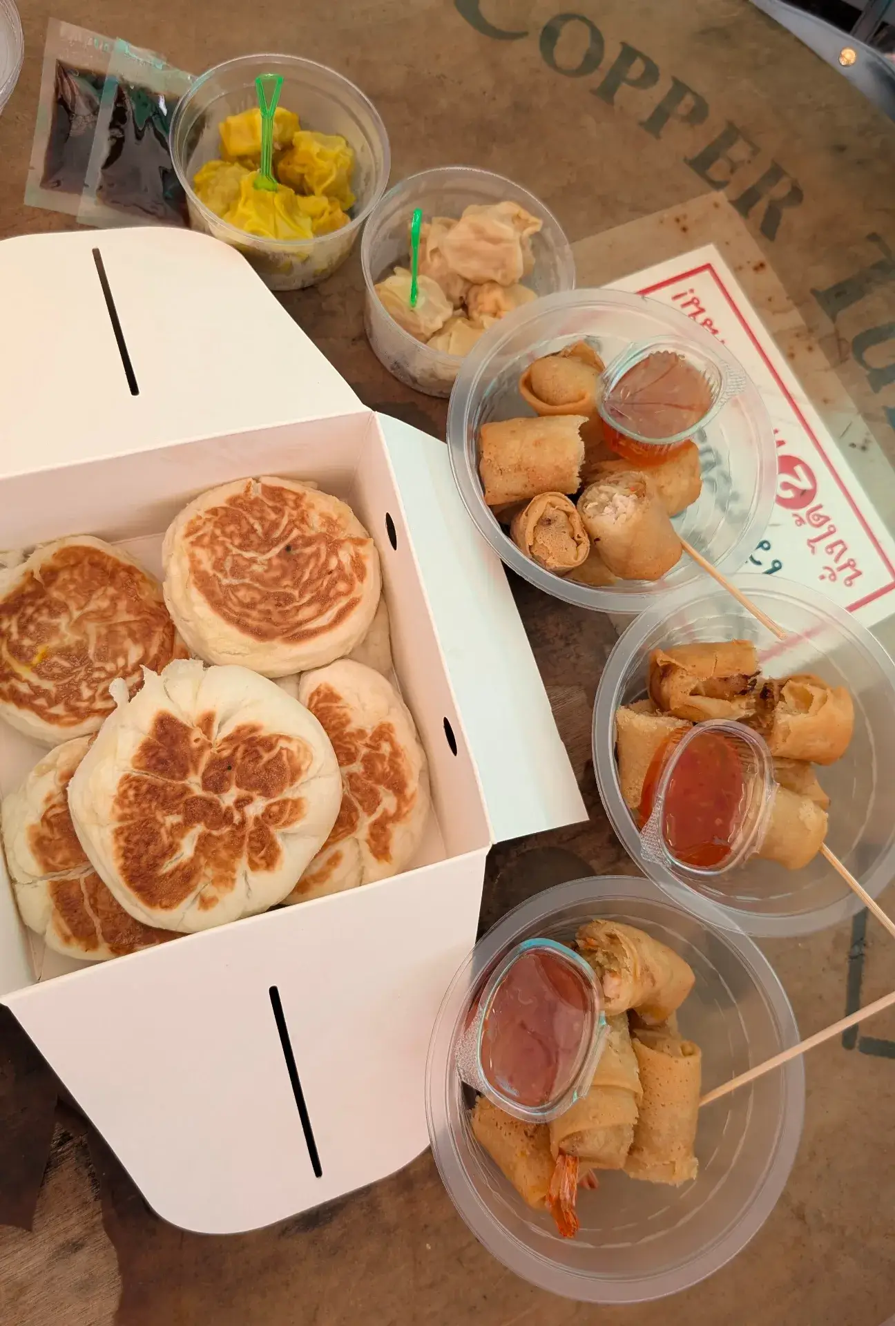 活気あるマーケットでテイクアウトした、タイの多彩なストリートフード。サクサクに揚げられた春巻き（ポピア・トード）にはスイートチリソースが添えられ、他にもシュウマイなどの点心や、こんがりと焼き目のついたおやき風のパンが並びます。手軽につまめるこれらの軽食は、観光の合間のエネルギー補給にぴったりな、旅の日常を彩る美味しい一コマです。