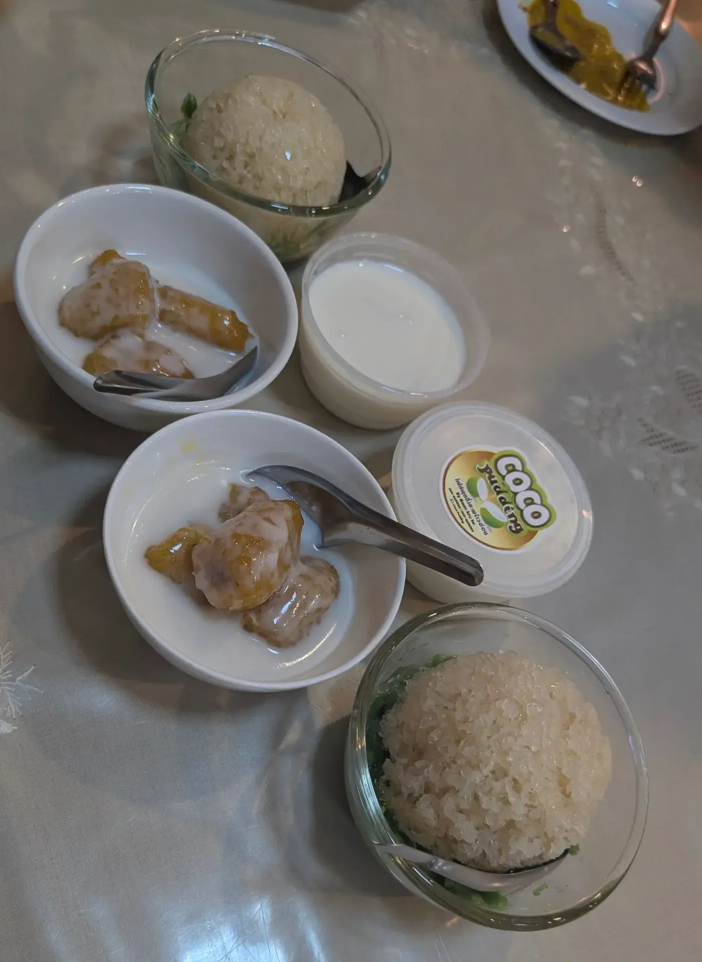 タイ料理のディナーを締めくくる、バラエティ豊かな伝統スイーツ。ココナッツミルクをかけた甘いモチ米（カオニャオ・マムアン風）や、温かいバナナのシロップ煮、さらに「COCO PUDDING」と書かれたカップ入りのココナッツプリンなど、現地の素材を活かした素朴で優しい甘みのデザートセットです。