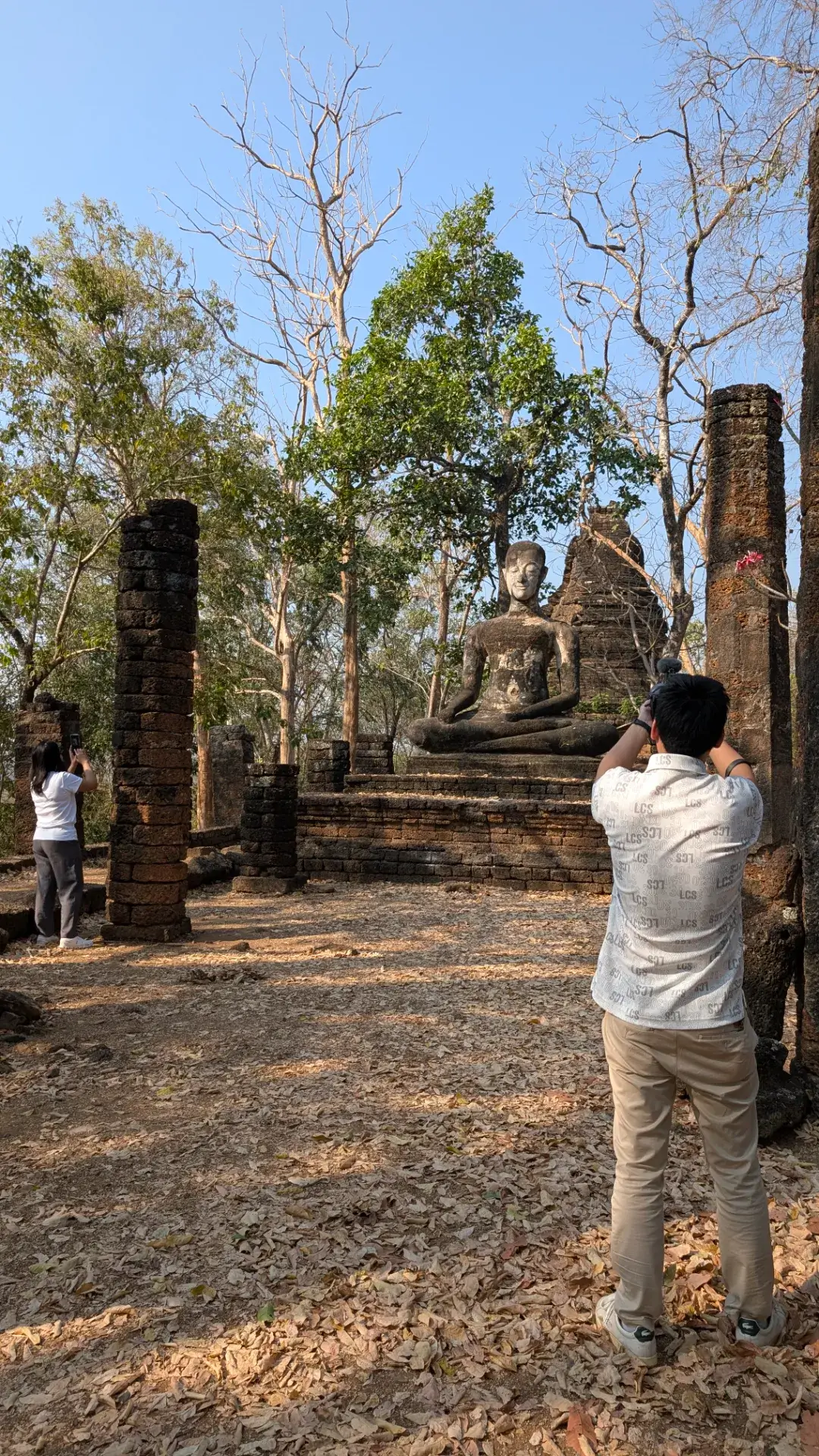 タイのシーサッチャナーライ歴史公園にて、レンガ造りの柱の間に座る仏像を背後から撮影する男性。左側にはスマートフォンのカメラを構える女性の姿も見え、歴史的な遺構と現代の旅人が交差する風景。