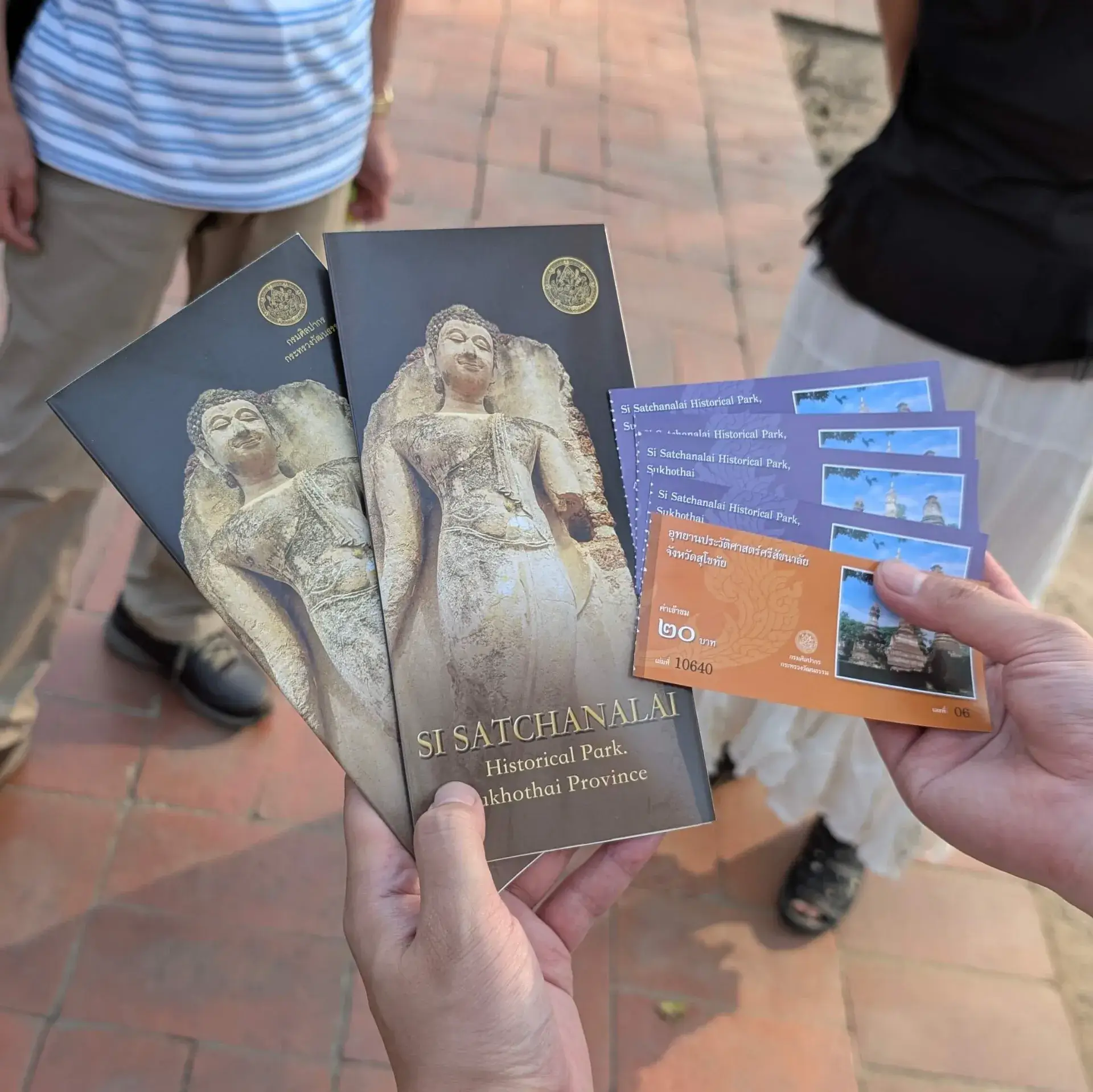 タイのスコータイ県にあるシーサッチャナーライ歴史公園の入場チケットとパンフレットを持つ手。パンフレットには美しい仏像が描かれ、背景には旅の仲間たちの姿が見える、歴史探索の始まりを告げる一枚。