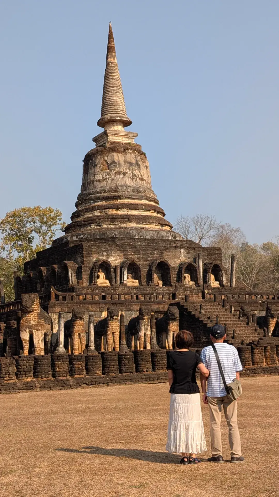 タイのシーサッチャナーライ歴史公園にある「ワット・チャーンローム」。基壇を39頭の象の彫刻が囲む巨大な鐘型仏塔（チェディ）と、その前で寄り添って遺跡を見上げる男女の後ろ姿。青空の下に広がる歴史的景観。
