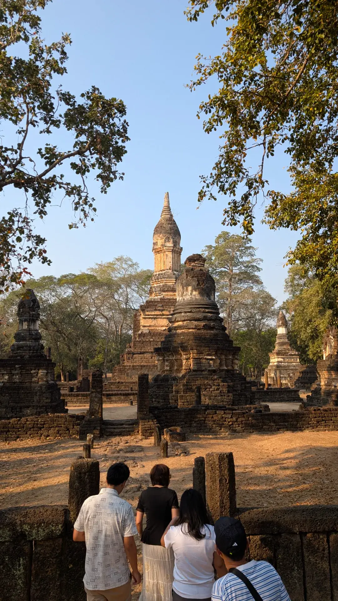 タイのシーサッチャナーライ歴史公園にある「ワット・チェーディー・チェット・テーオ」。朝の光を浴びてそびえ立つ複数の仏塔（チェディ）と、それを見つめる旅の仲間たちの後ろ姿。スコータイ王朝の歴史を感じさせる静謐な風景。